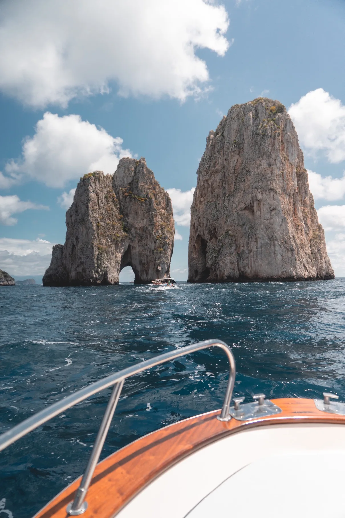 Boat bow pointing toward two towering sea stacks with a natural arch rising from deep blue waters under a partly cloudy sky