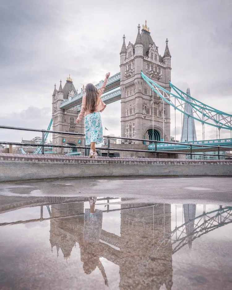 Woman in a floral skirt and pink jacket waving toward Tower Bridge in London, with her reflection visible in a puddle in the foreground.