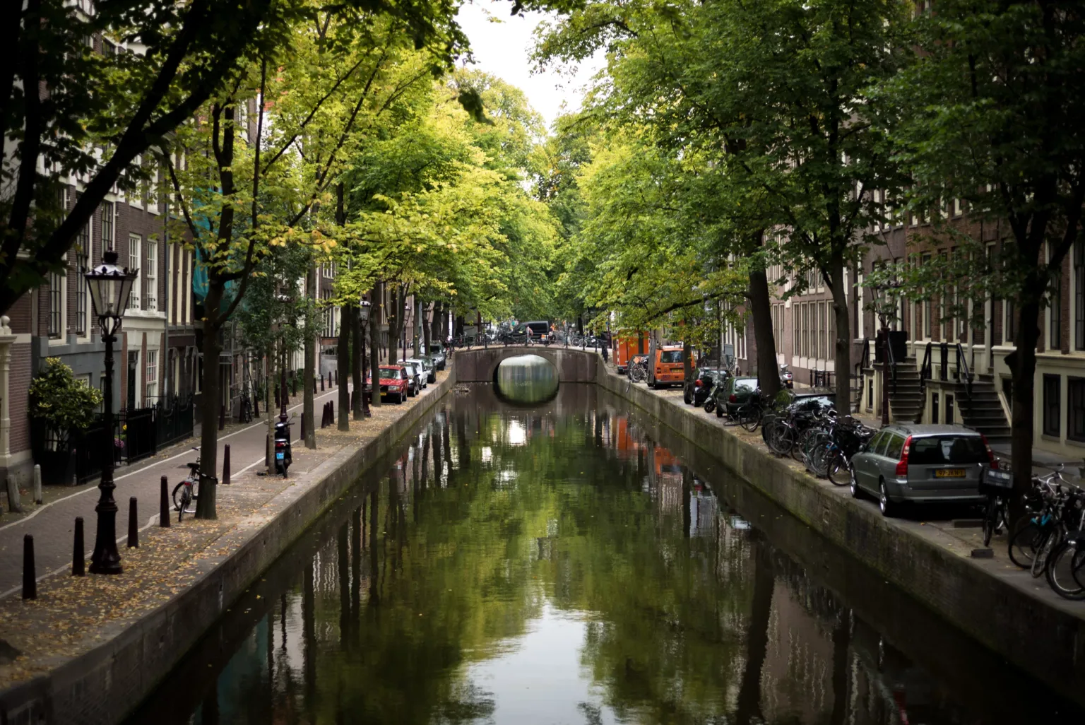 Tree-lined canal flanked by narrow townhouses, parked bikes and cars with a small arched bridge in the distance.