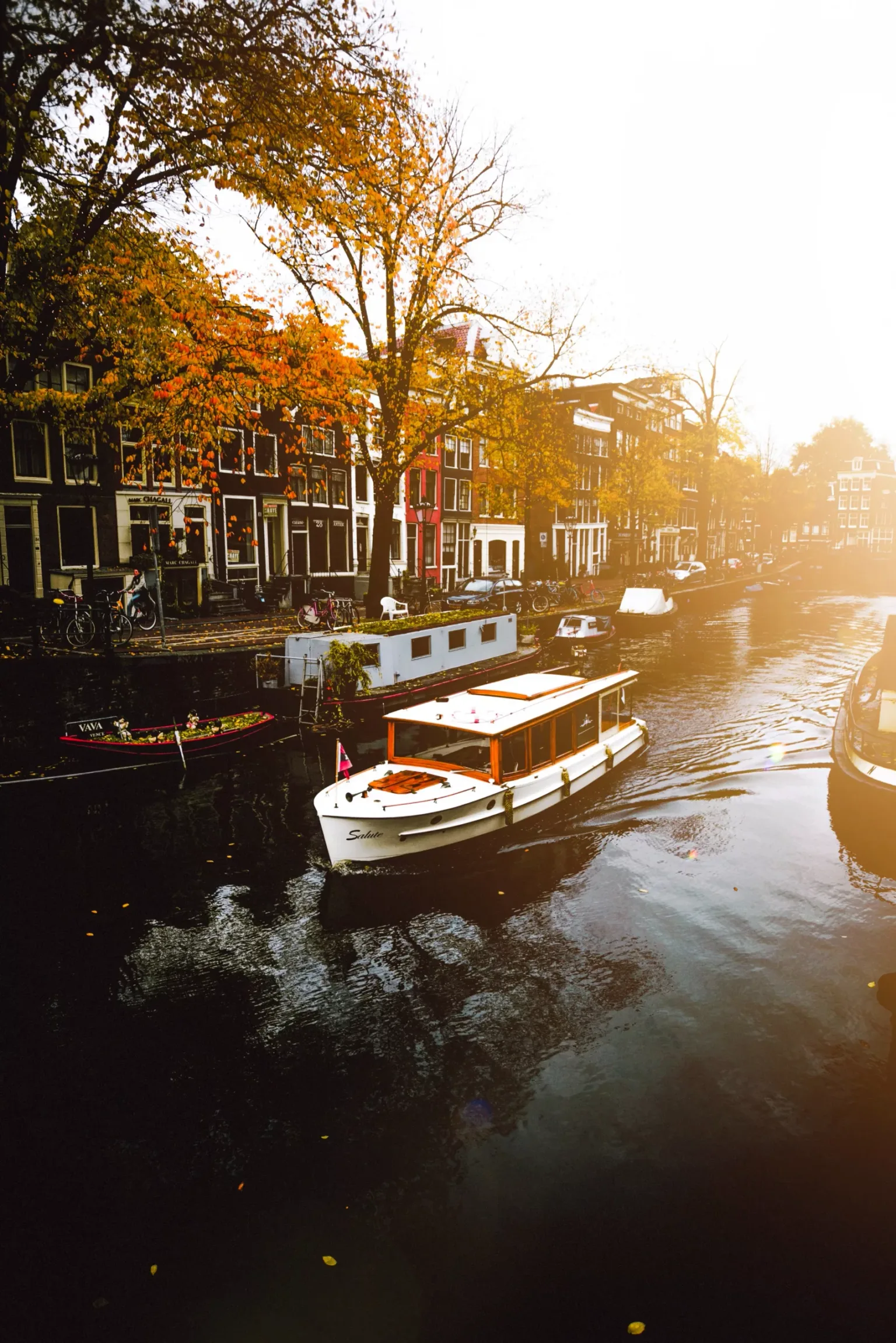 Canal boat gliding past autumn trees and tall narrow townhouses along a sunlit European canal.
