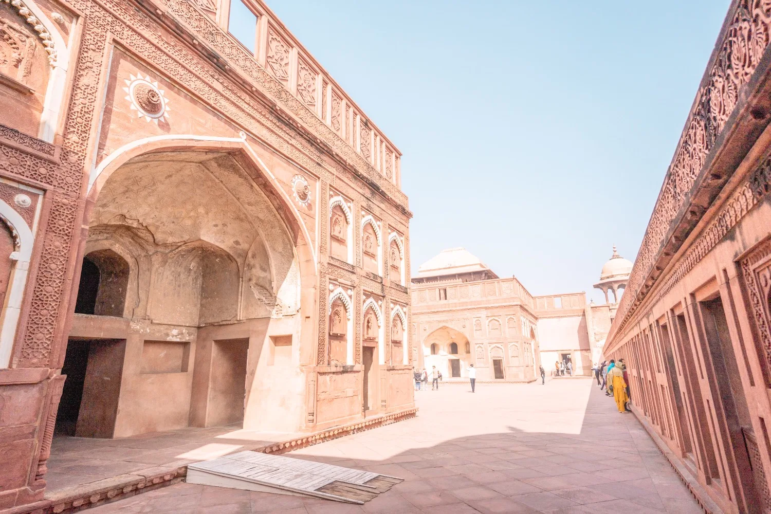 Sunlit courtyard of a red sandstone Mughal palace with ornate arches and visitors exploring the site