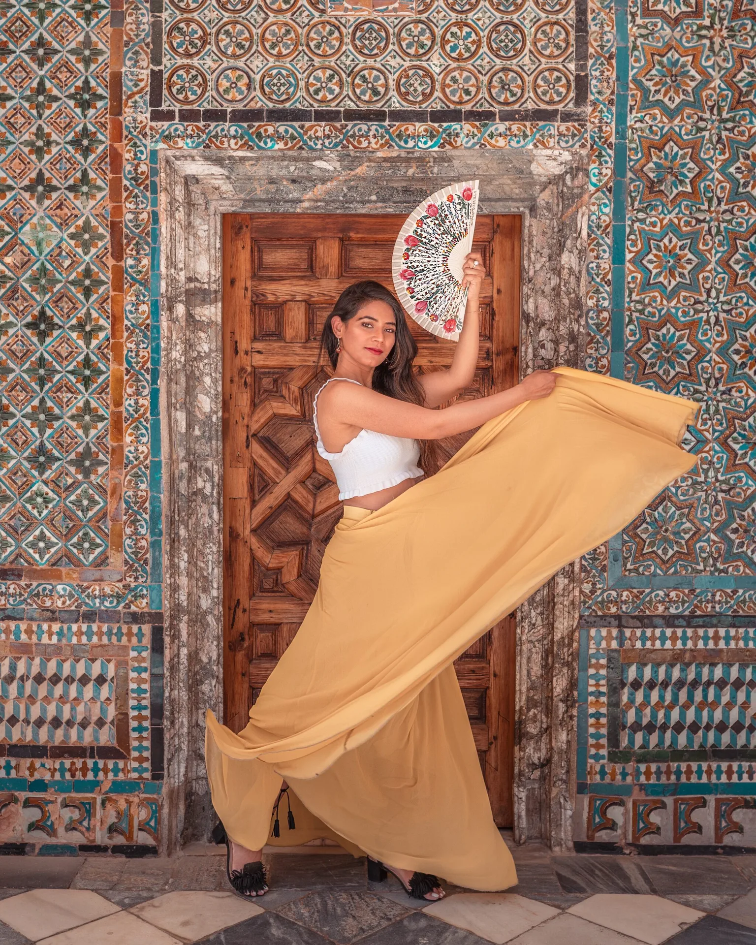 Woman in a flowing mustard skirt and white top poses with a decorative hand fan in front of a carved wooden door framed by intricate blue and orange mosaic tiles.