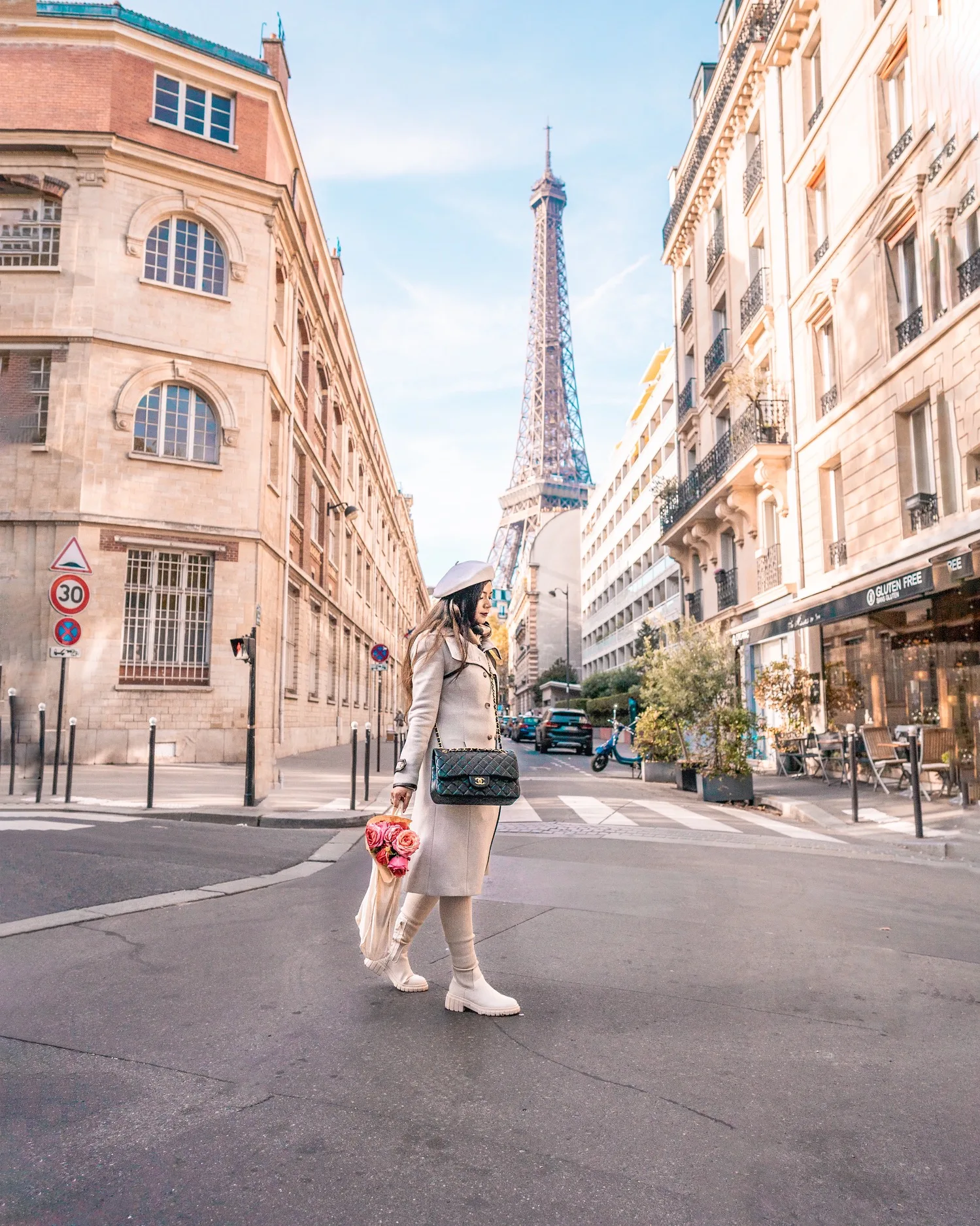 Woman in a cream coat and beret crossing a Paris street with a bouquet and the Eiffel Tower rising between Haussmannian buildings behind her.