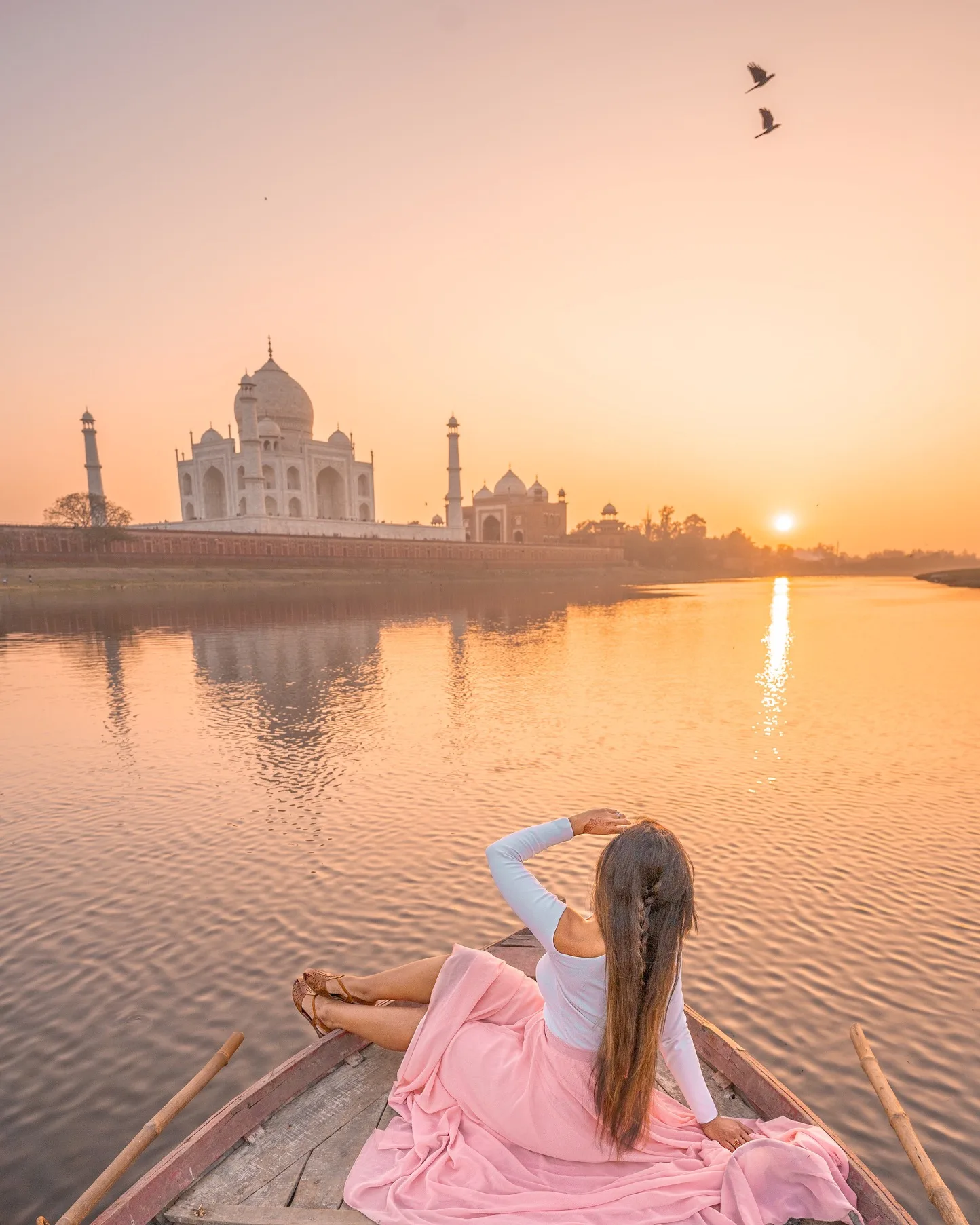Woman in a pink skirt sitting at the bow of a boat watching sunrise over the Taj Mahal reflected in the river