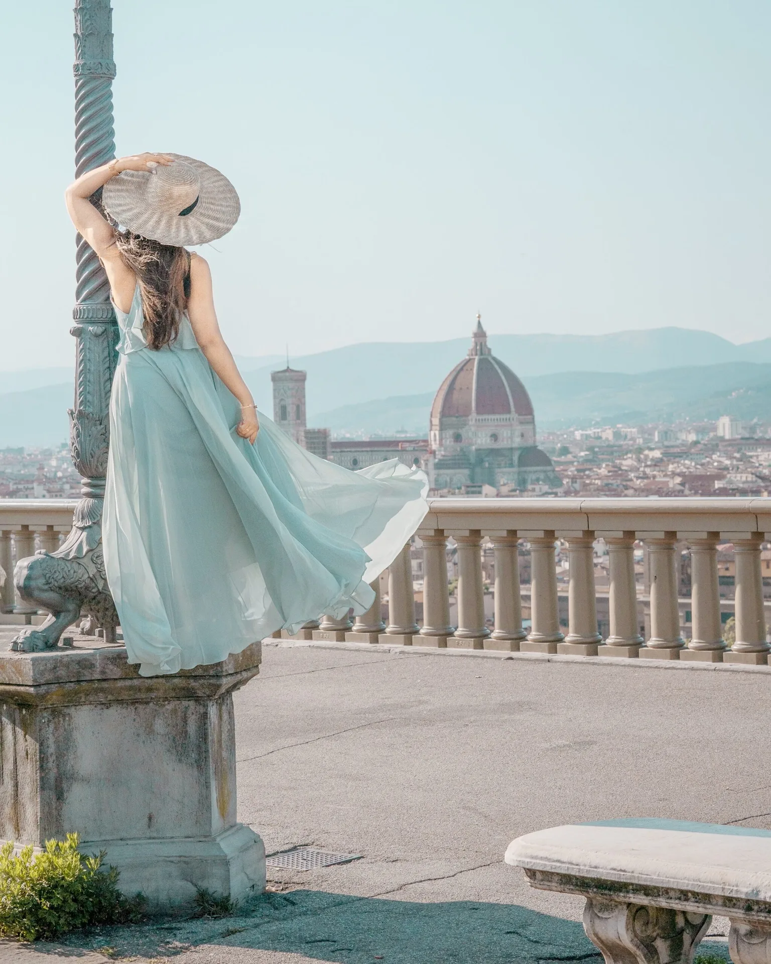 Woman in a flowing pale blue dress and wide-brimmed hat gazes over Florence with the Duomo visible across the city.
