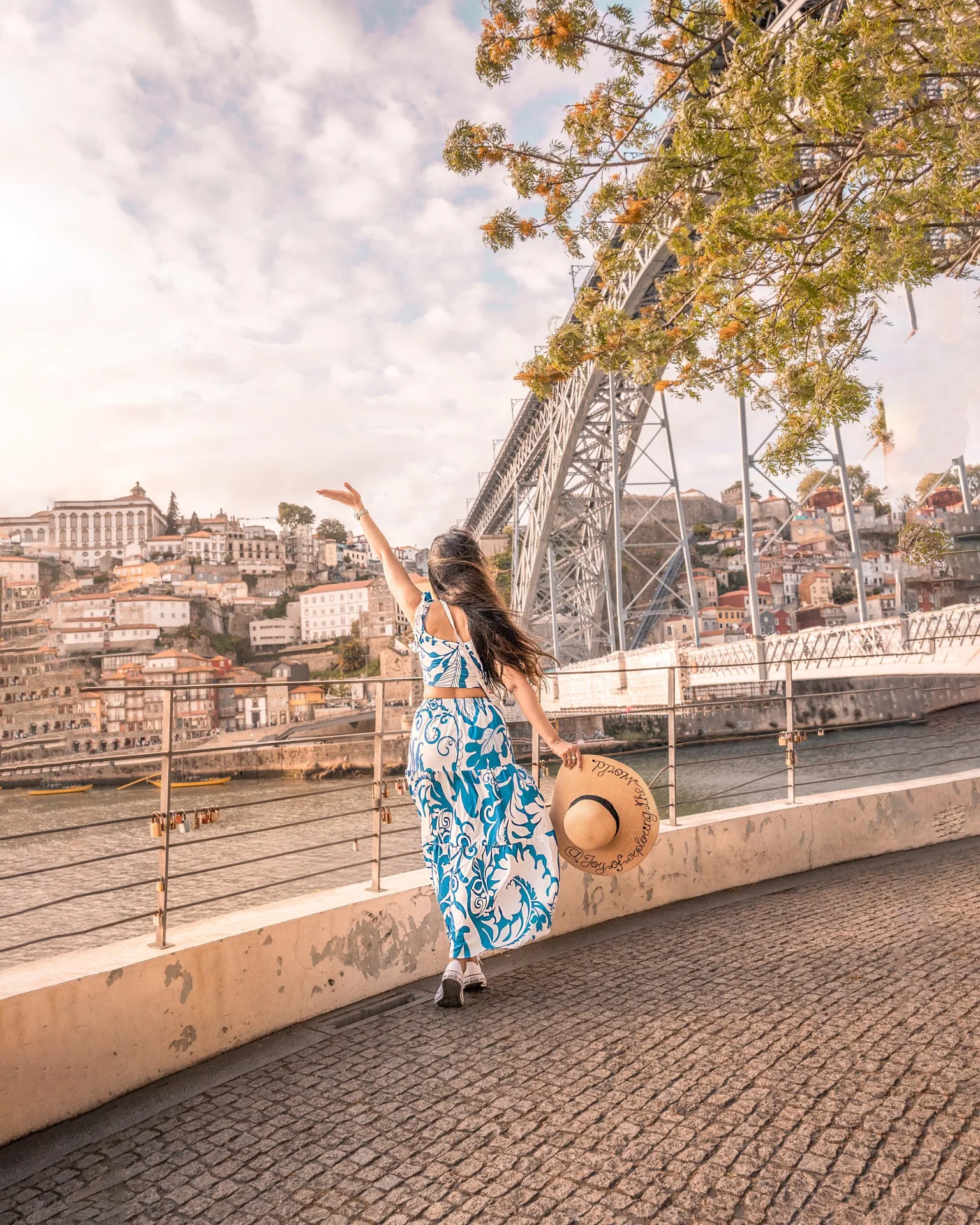Woman in a blue patterned dress holding a sunhat and waving toward the riverfront with the arched metal bridge and terraced city in the background.