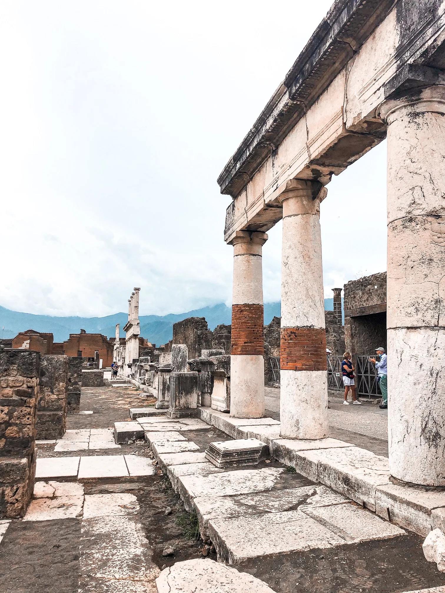 Ancient stone colonnade and ruins under a pale sky with distant mountains, visited by a few tourists.