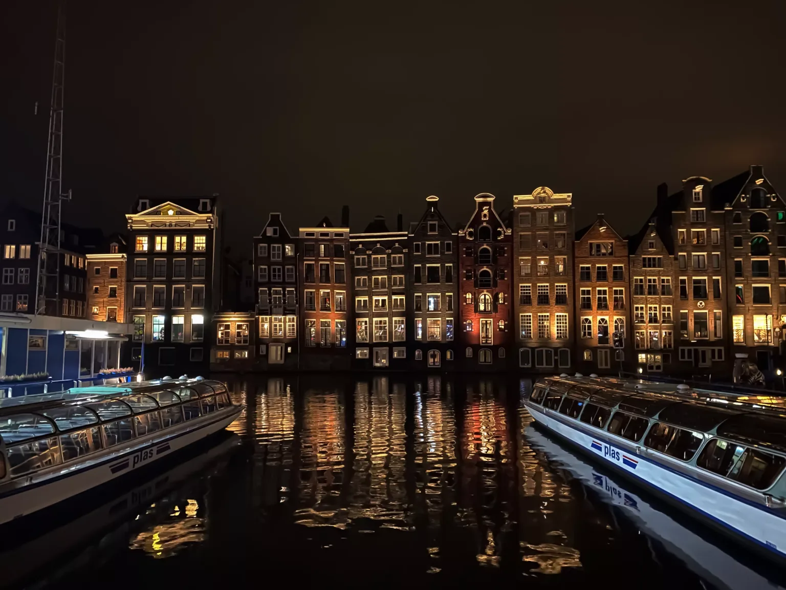 Canal at night with illuminated narrow Dutch gabled houses reflected in the water and glass-roofed tour boats docked either side.