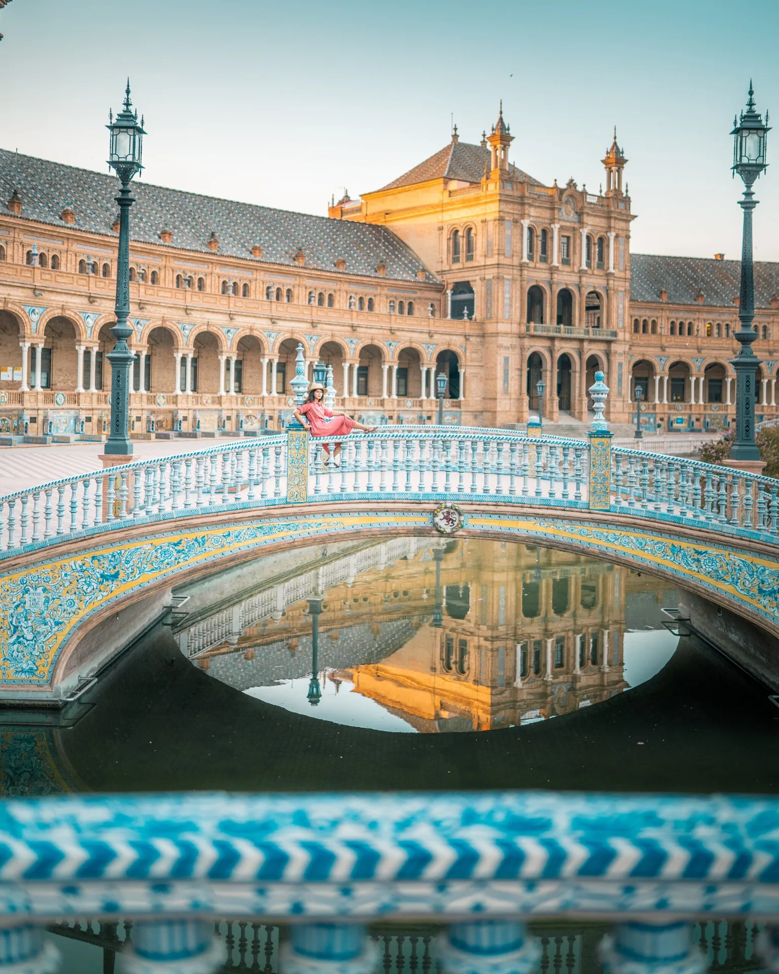 Woman in a pink dress lounging on a decorative tiled bridge at the historic Plaza de España with the building reflected in the canal.