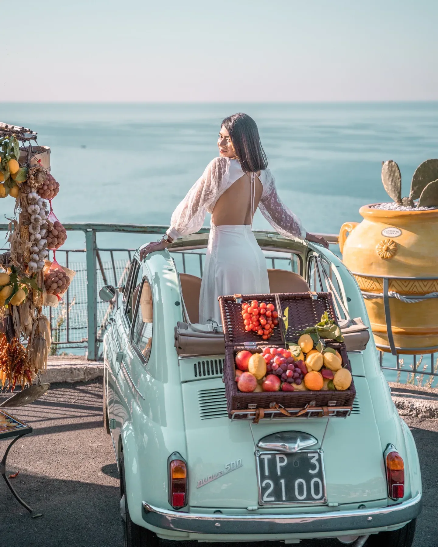 Best Instagram spots Amalfi Coast - Woman in a white backless dress standing in a vintage pale-green Fiat 500 with a picnic basket of fruit, overlooking a sunlit Mediterranean sea.