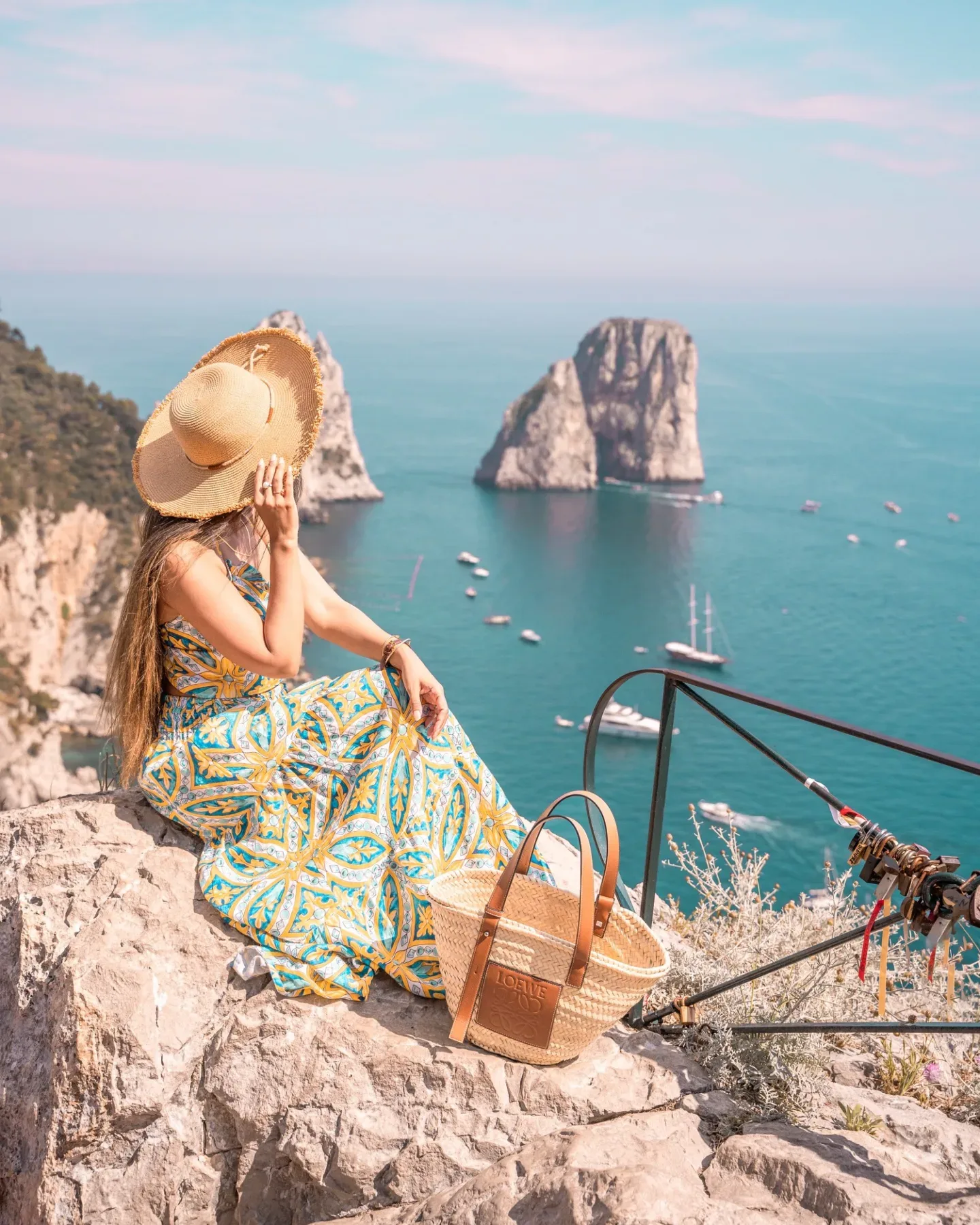 Woman in a patterned sundress and wide straw hat sitting on a cliff overlooking turquoise sea and rocky islets, with a woven tote beside her.