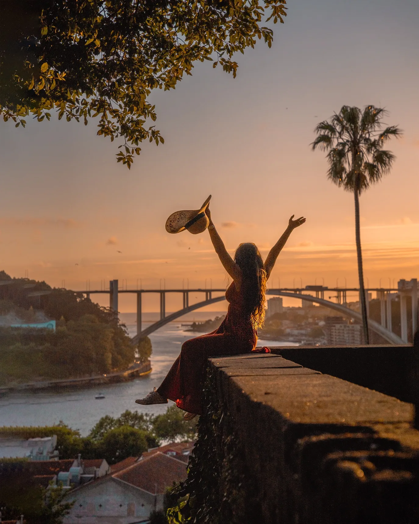 Woman sitting on a stone wall at sunset with arms raised, holding a hat, overlooking a river and arched bridge.