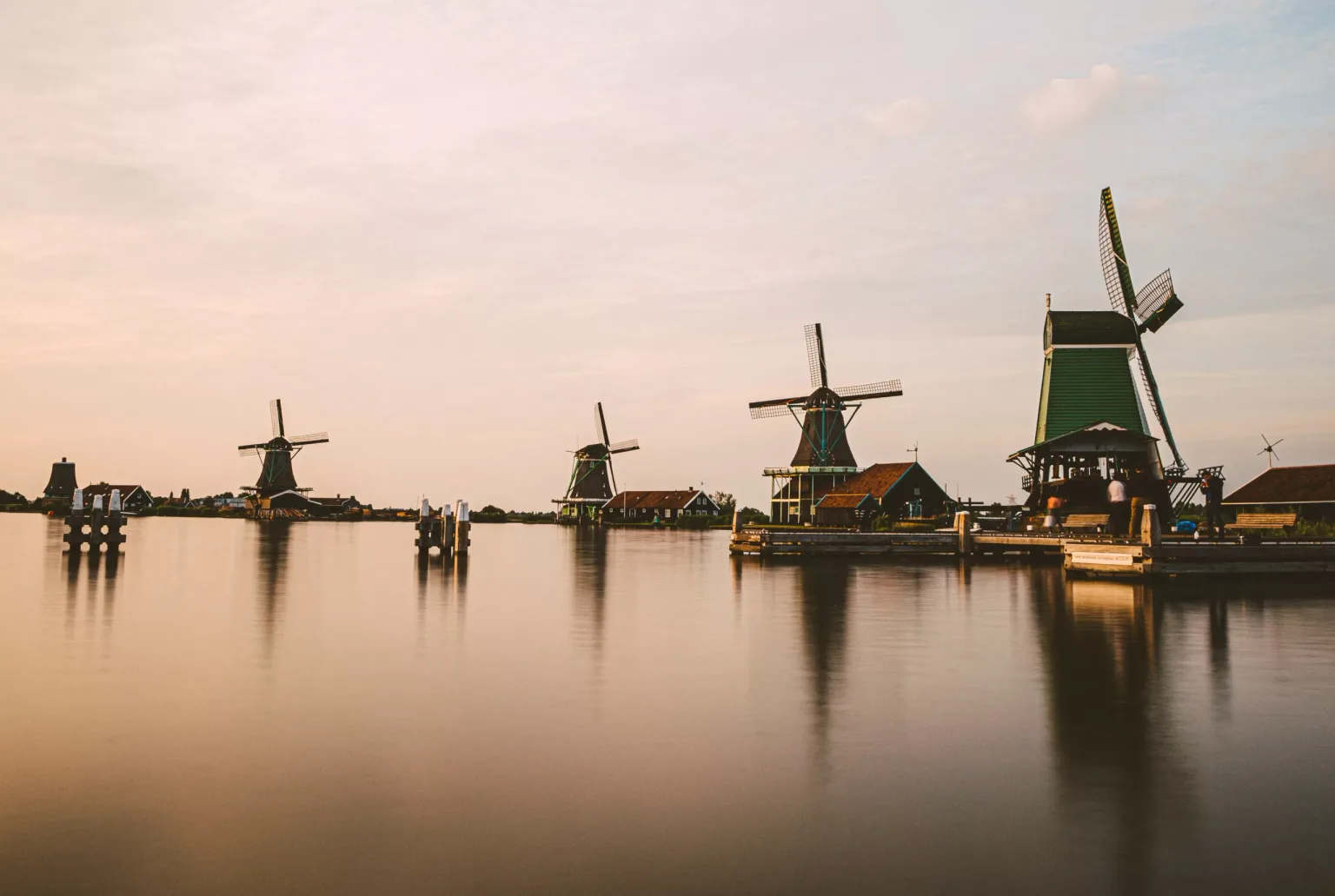 Row of traditional Dutch windmills reflecting on calm water at sunset with a small dock in the foreground.