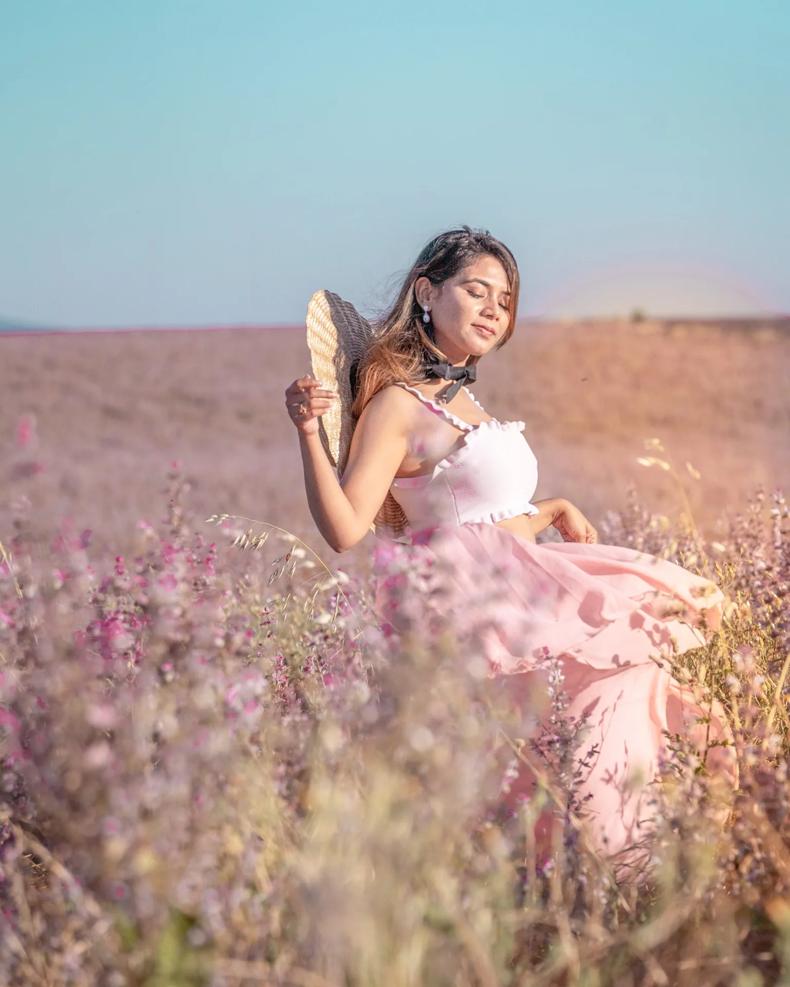 Woman in a light pink dress holding a straw hat while standing in a sunlit wildflower field with eyes closed.