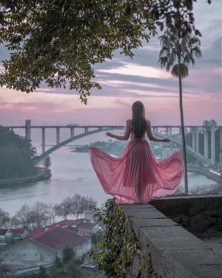 Woman in a flowing pink dress stands on a stone ledge overlooking a river and arched bridge at sunset.
