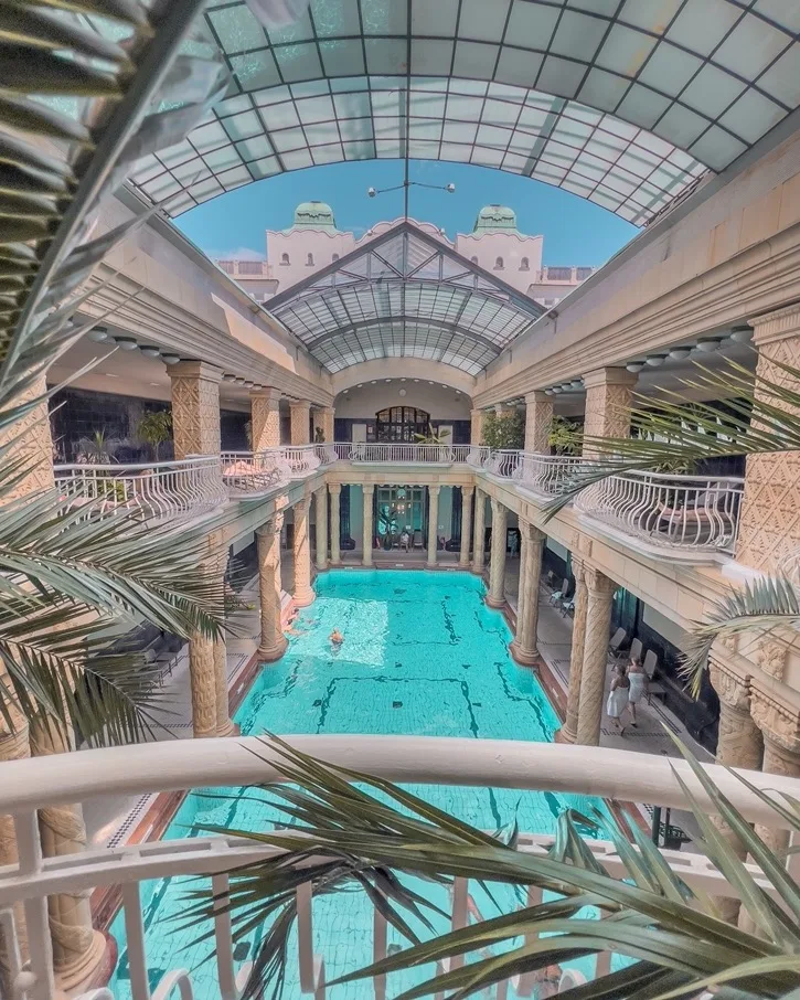Sunlit indoor turquoise pool surrounded by classical columns and balconies beneath a glass arched roof, framed by palm fronds.
