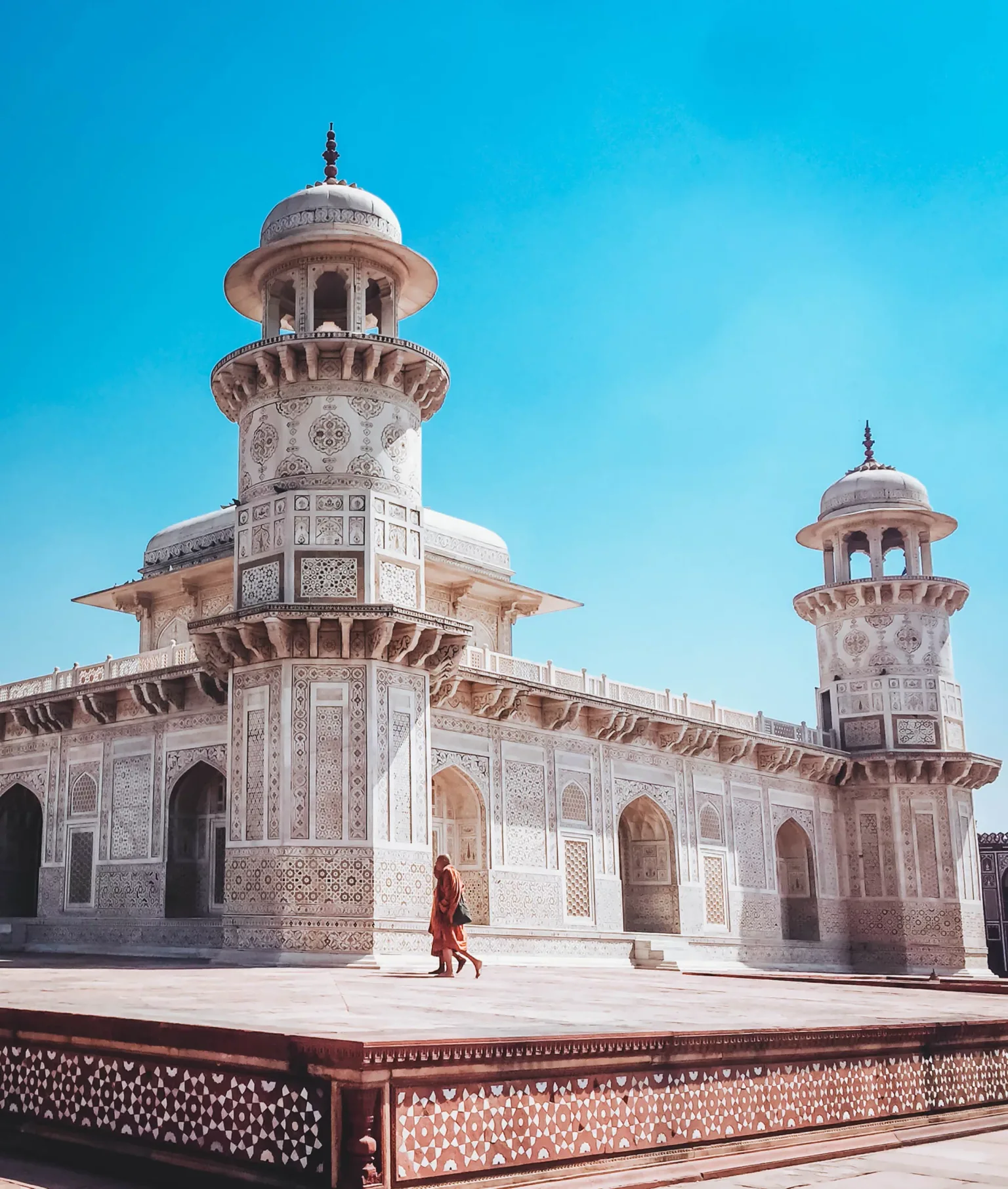 Ornately carved white marble mausoleum with two domed towers under a bright blue sky and a lone visitor walking on the courtyard.