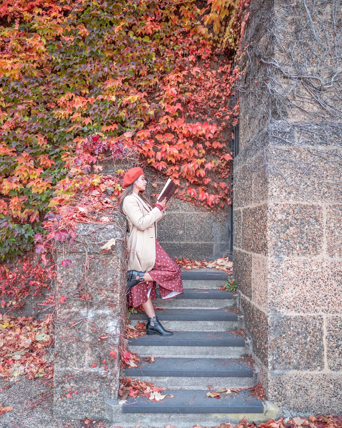 Woman in a red beret and beige coat reading a book on stone steps surrounded by vibrant autumn ivy and fallen leaves