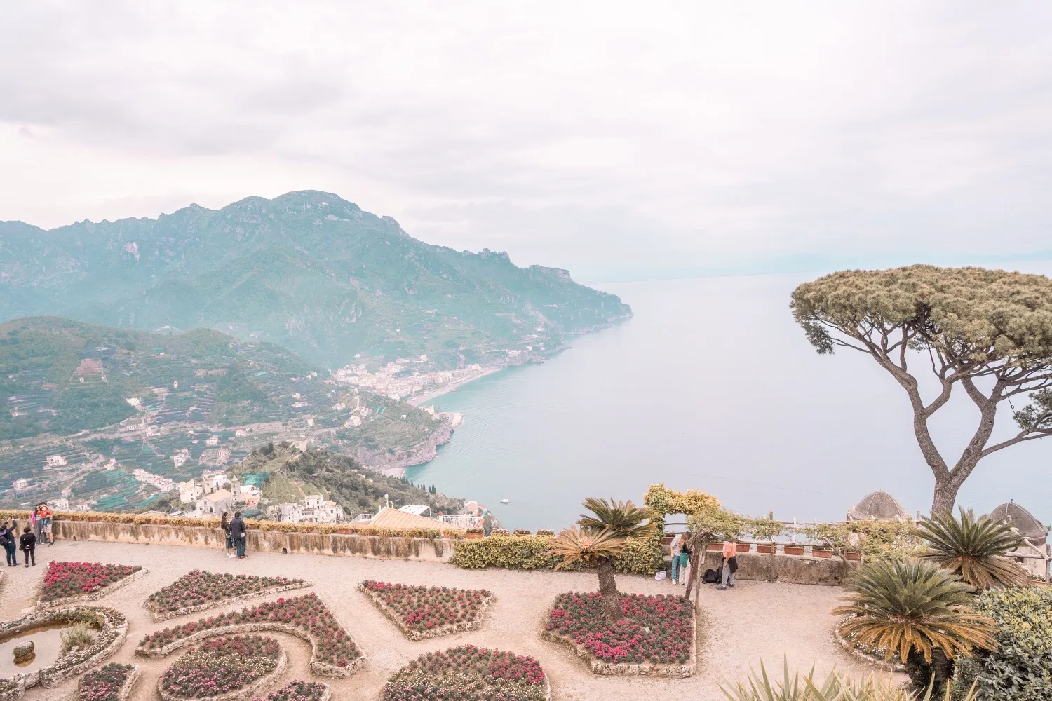 Terraced flower gardens and stone balustrade overlooking a coastal cliff, Mediterranean sea and distant mountains under a pale sky.