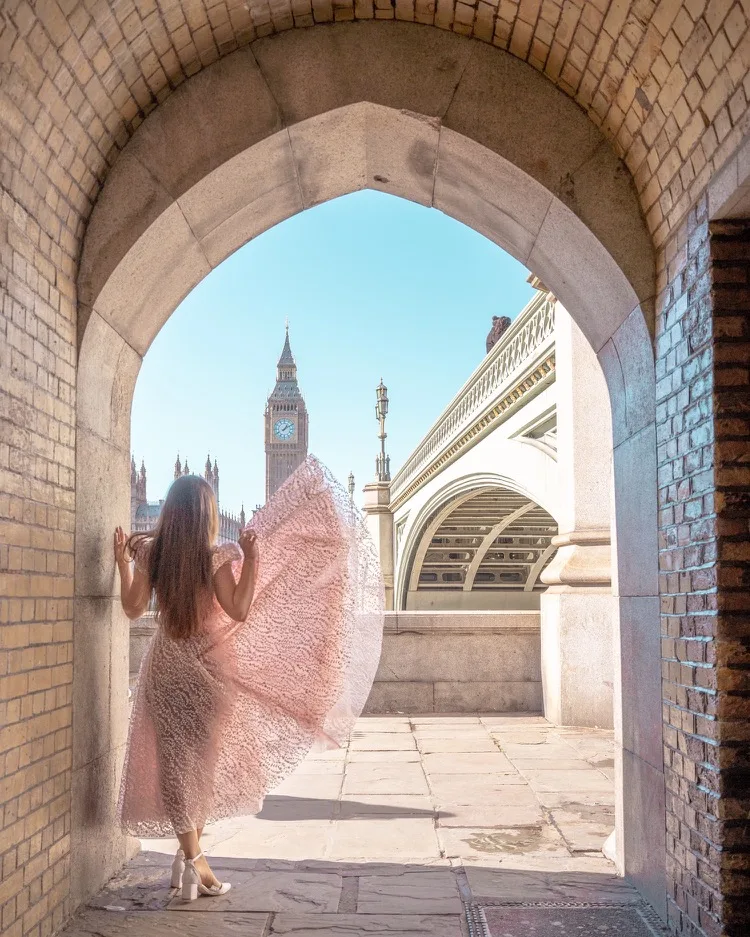 Woman in a flowing pink dress framed by a stone archway overlooking Big Ben and Westminster Bridge in London.