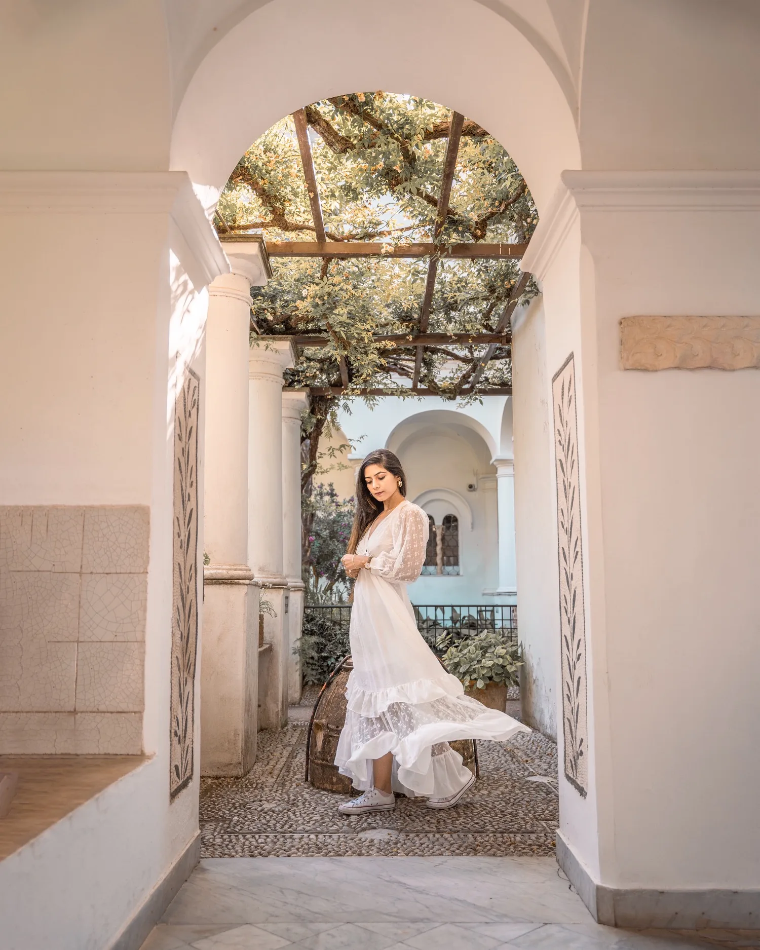 Young woman in a flowing white dress and sneakers stands beneath an arched, vine-covered courtyard walkway, sunlight filtering through the trellis.