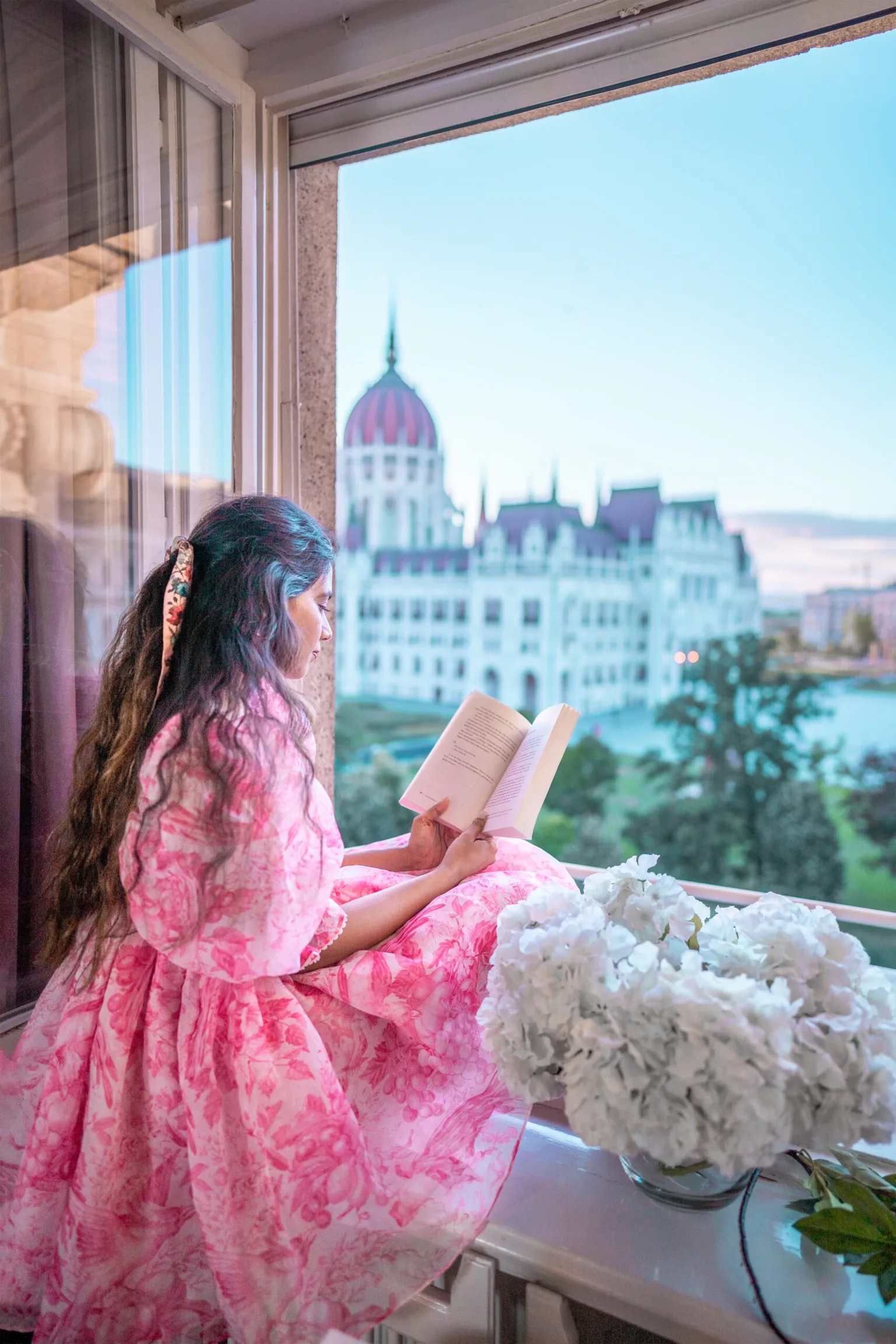 Woman in a pink floral dress reading a book on a windowsill overlooking the domed Parliament building and river at dusk