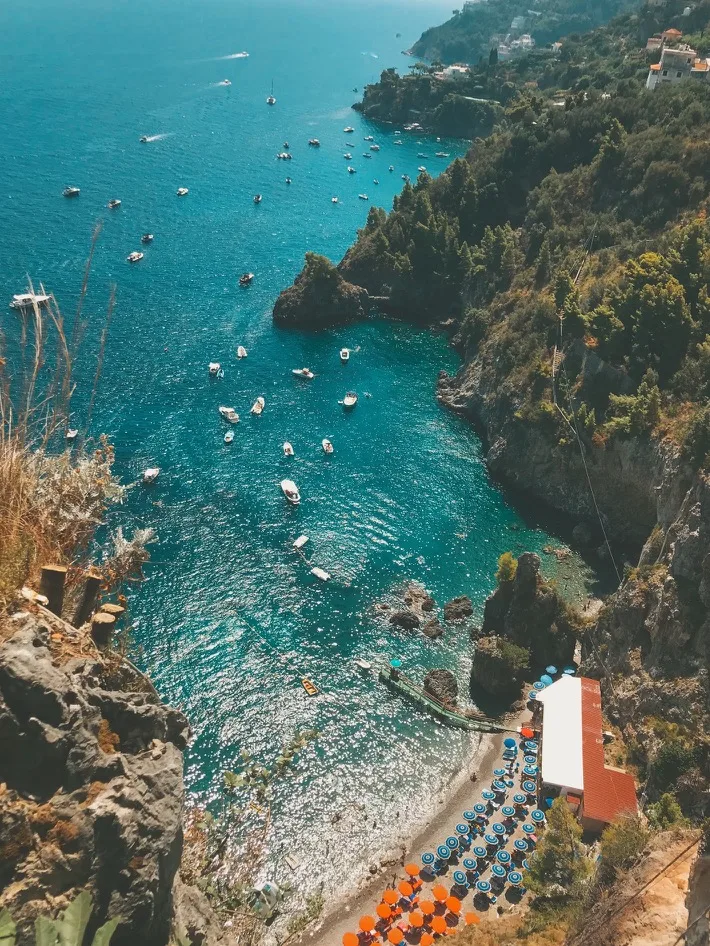 Clifftop view down to a turquoise cove dotted with boats and a pebble beach lined with colourful umbrellas.
