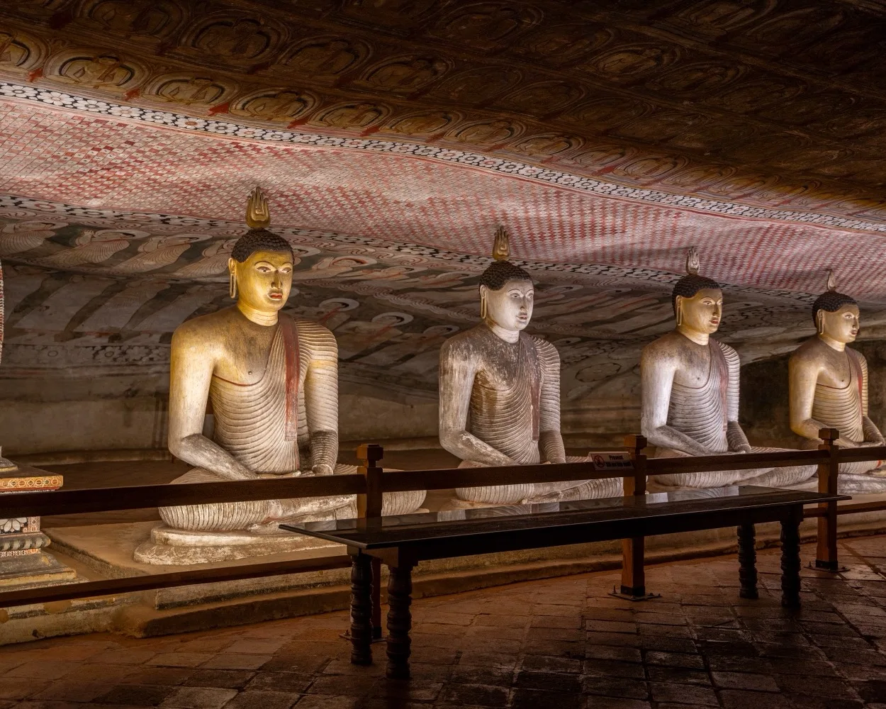 Long row of seated, meditative Buddha statues inside a dimly lit, ornately painted cave temple.