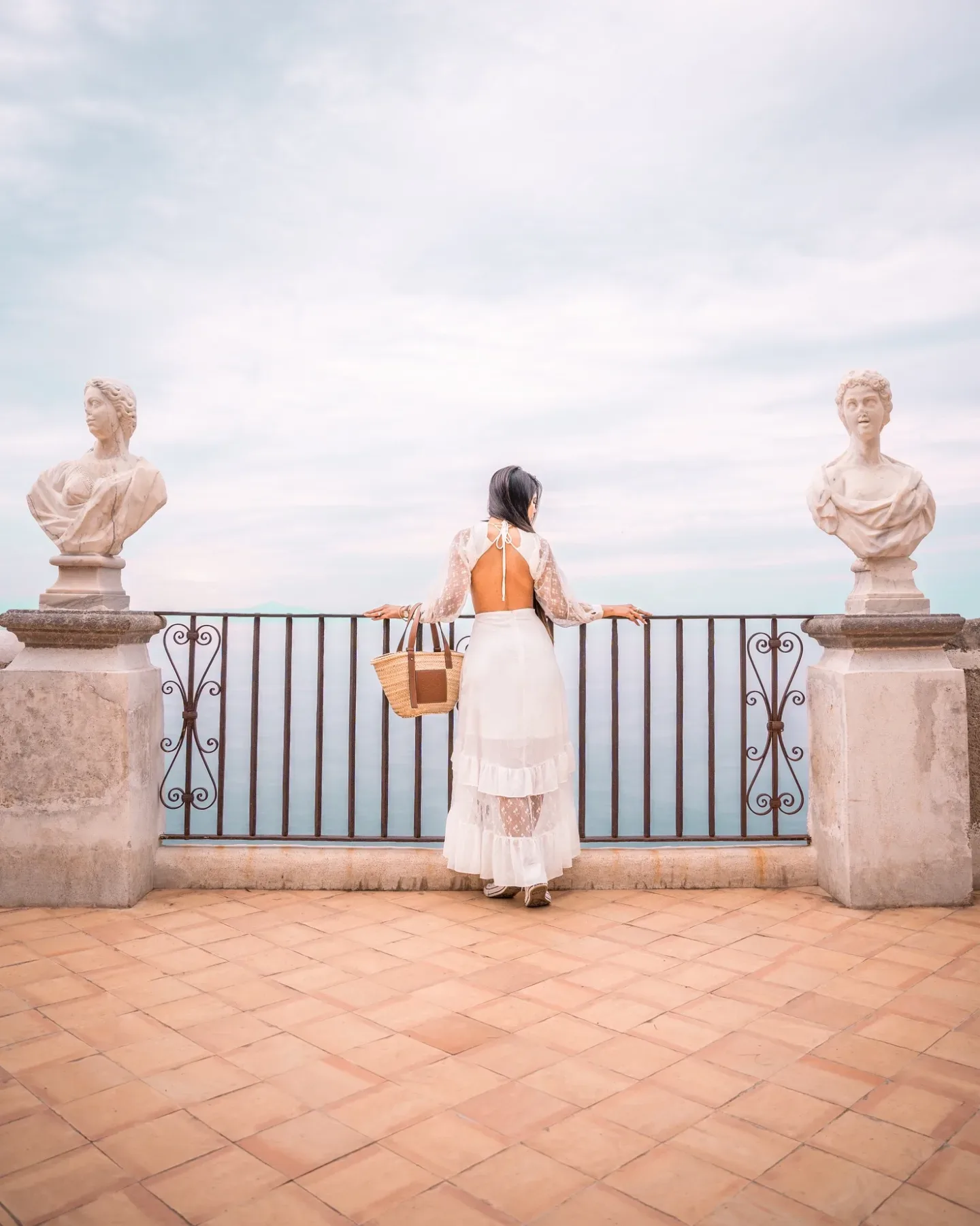 Woman in a white open-back dress holding a straw tote and leaning on a seaside terrace railing flanked by classical busts.