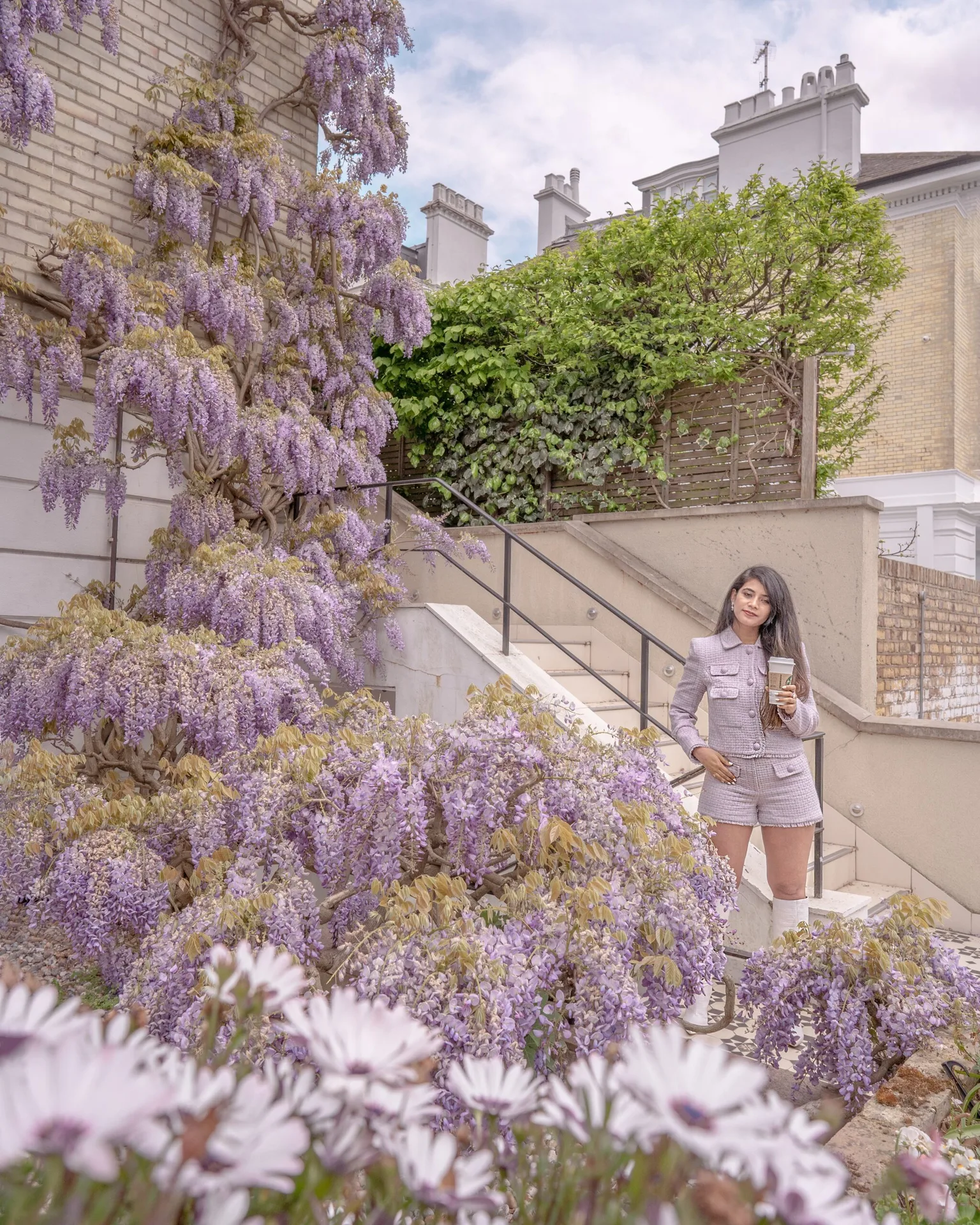 Woman in a lilac tweed co-ord holding a takeaway coffee beside a stairway, surrounded by cascading purple wisteria and spring blooms.