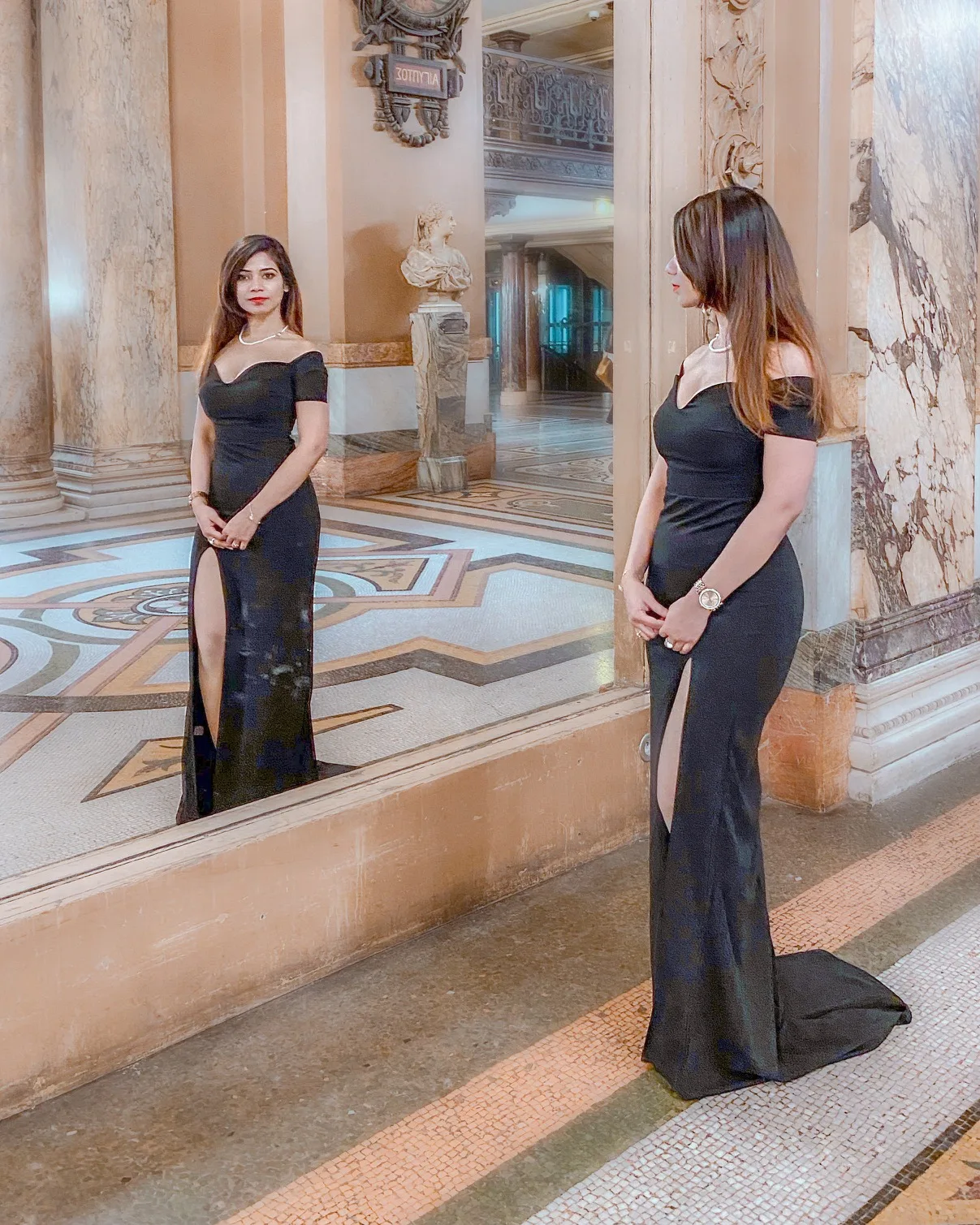 Woman in an elegant off-shoulder black gown with a thigh slit standing in a grand marble-floored hall, reflected in a large mirror.