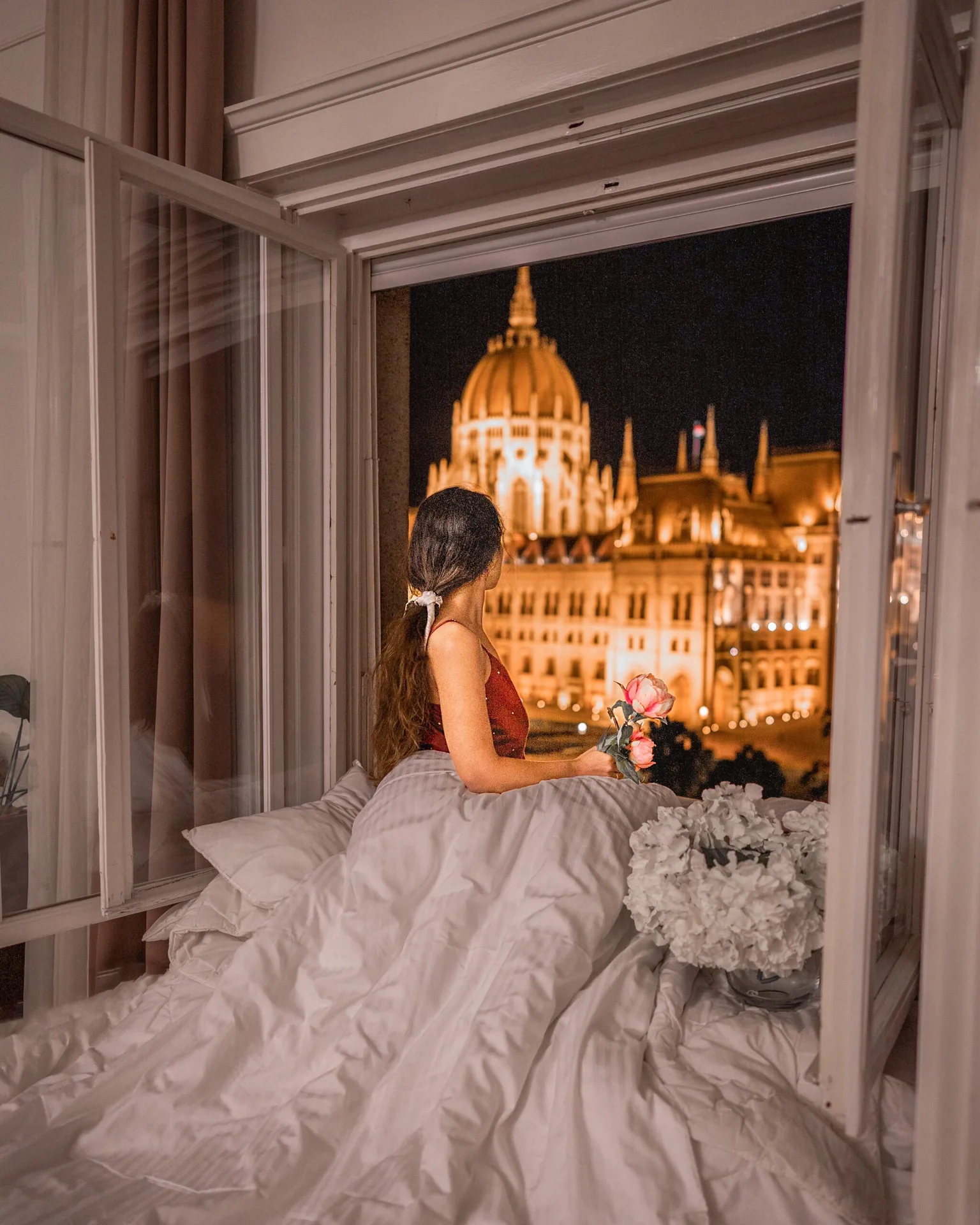 Woman in a hotel bed holding roses and gazing out an open window at a warmly lit domed landmark at night.