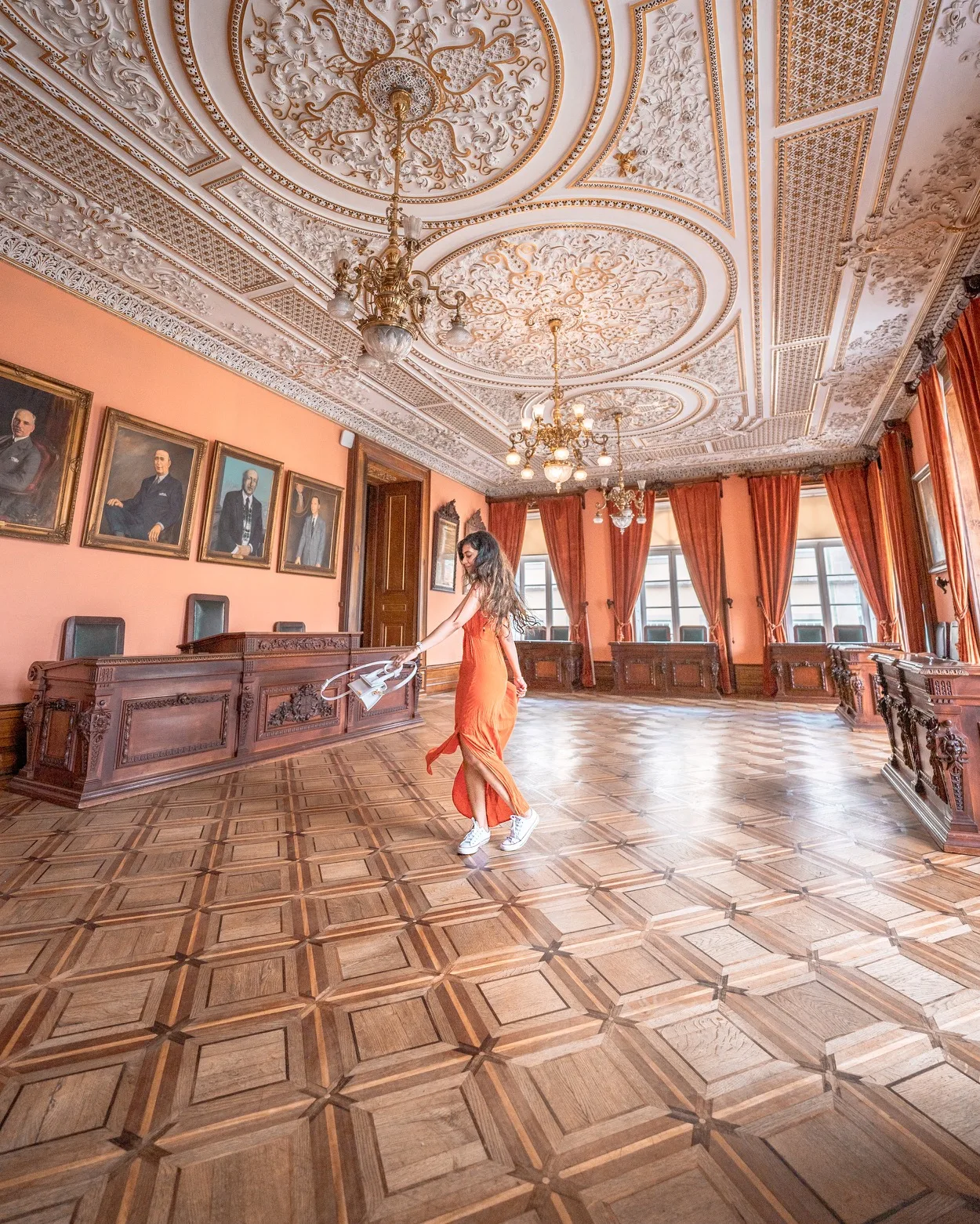 Woman in an orange dress twirling across an ornate parquet-floored hall with carved wooden benches, portraits and an elaborately moulded ceiling.