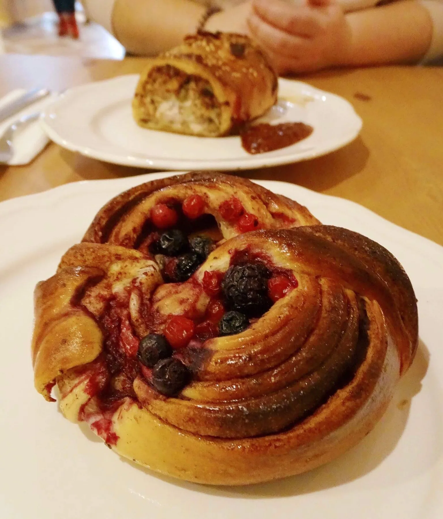 Sweet spiral bun filled with baked blueberries and red currants served on a white plate with another pastry in the background.
