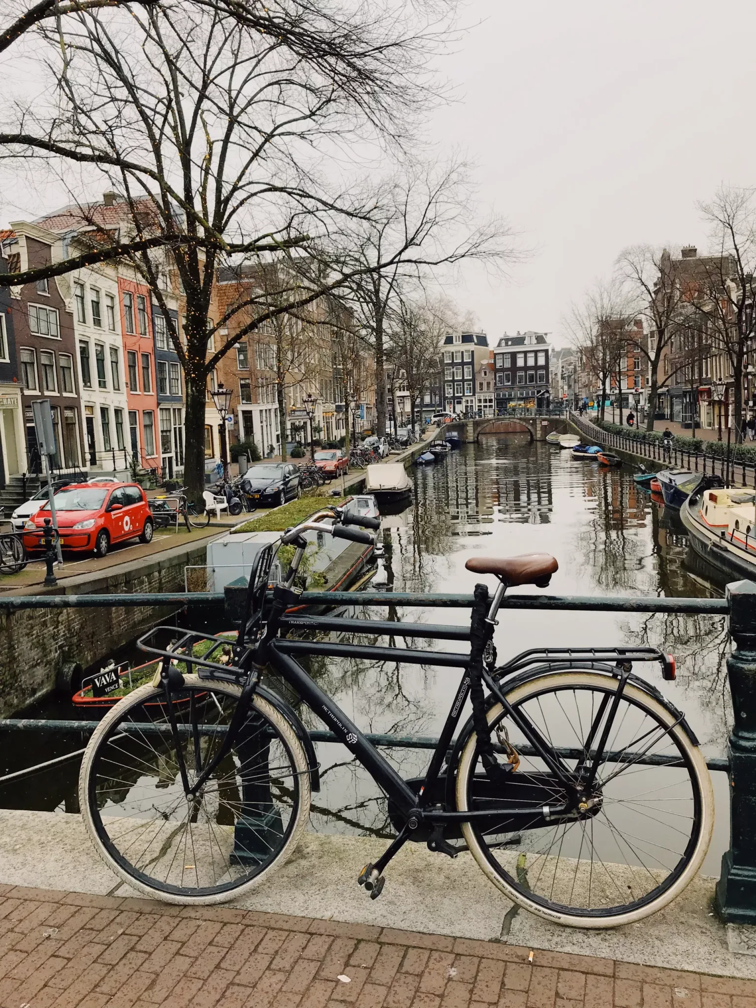 A black bicycle locked to a bridge railing overlooking a narrow Amsterdam canal lined with tall historic townhouses and moored boats.