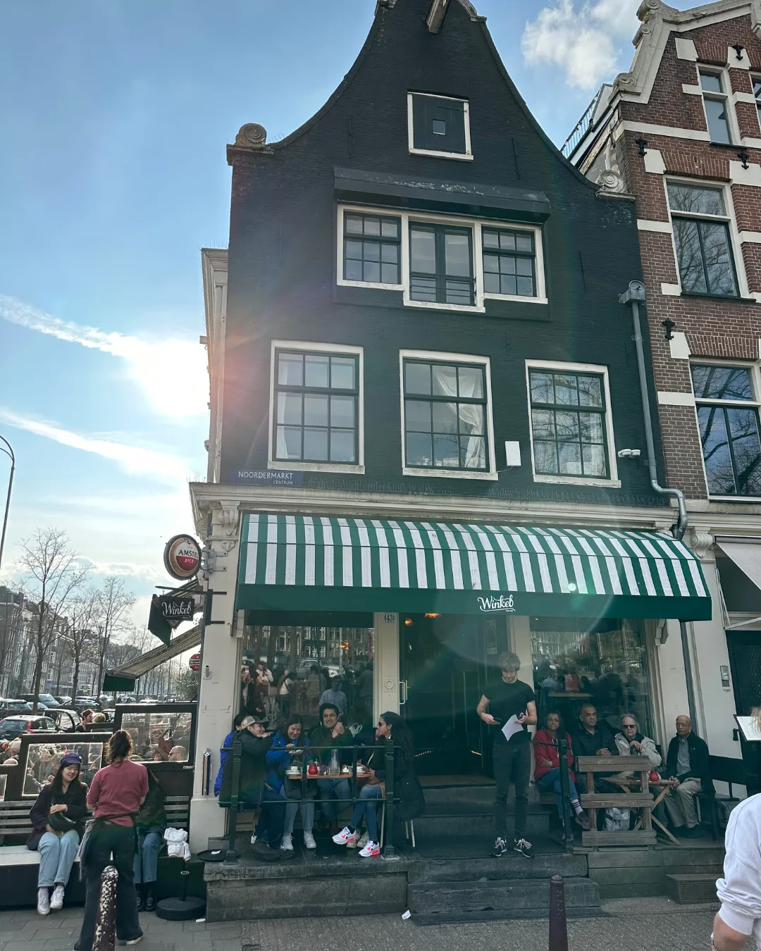 Historic narrow canal-house cafe with green-and-white striped awning and outdoor tables filled with people on a sunny day