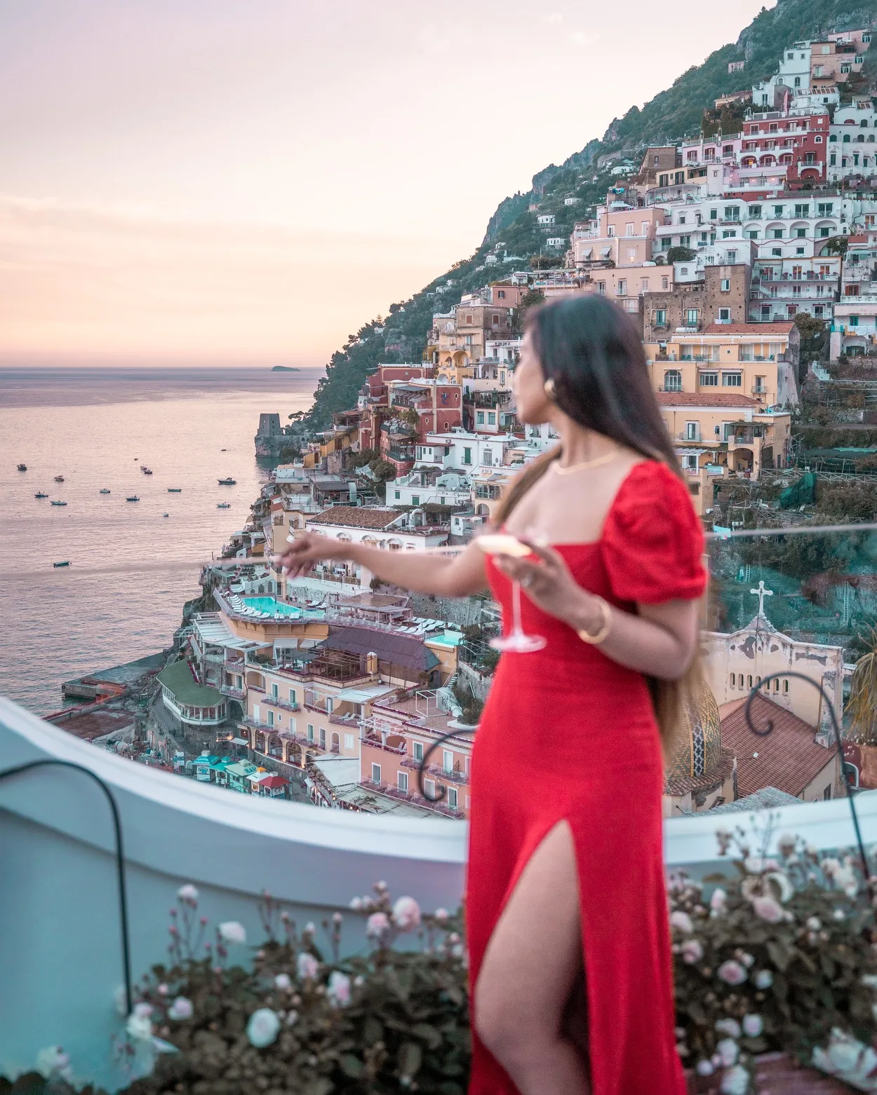 Best Things to do in Positano - Woman in a red dress holding a cocktail on a terrace overlooking pastel cliffside houses and the sea at sunset in Positano.