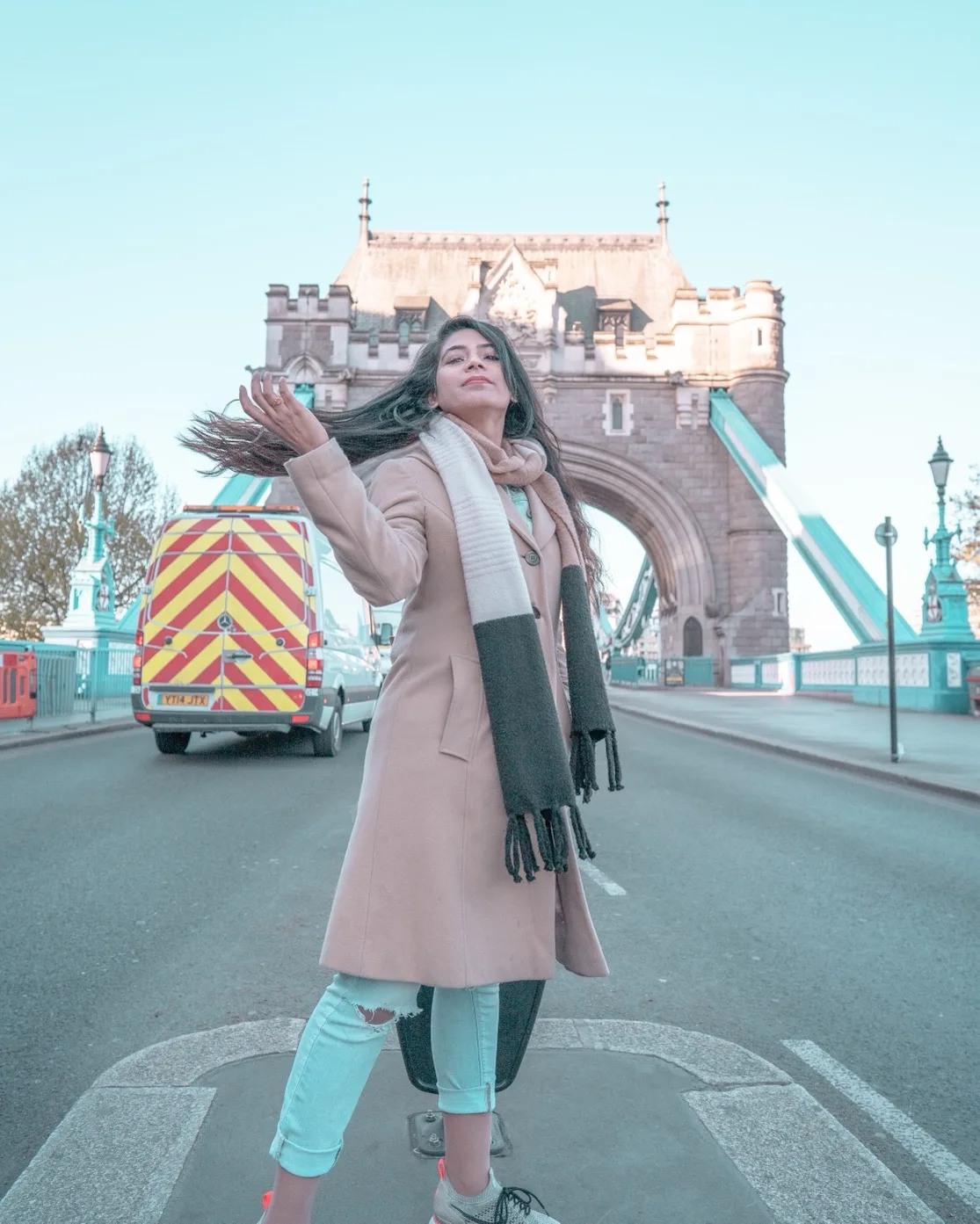Woman in a beige coat and long scarf flipping her hair while posing on Tower Bridge in London with a service van behind her
