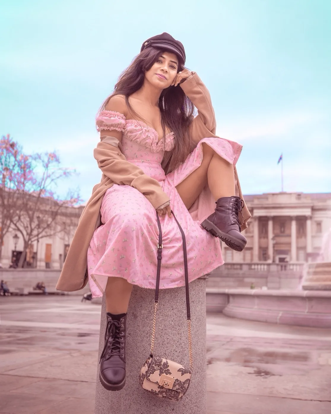 Woman in a pink off-shoulder dress, camel coat and black boots sitting on a stone bollard with a patterned handbag in a plaza