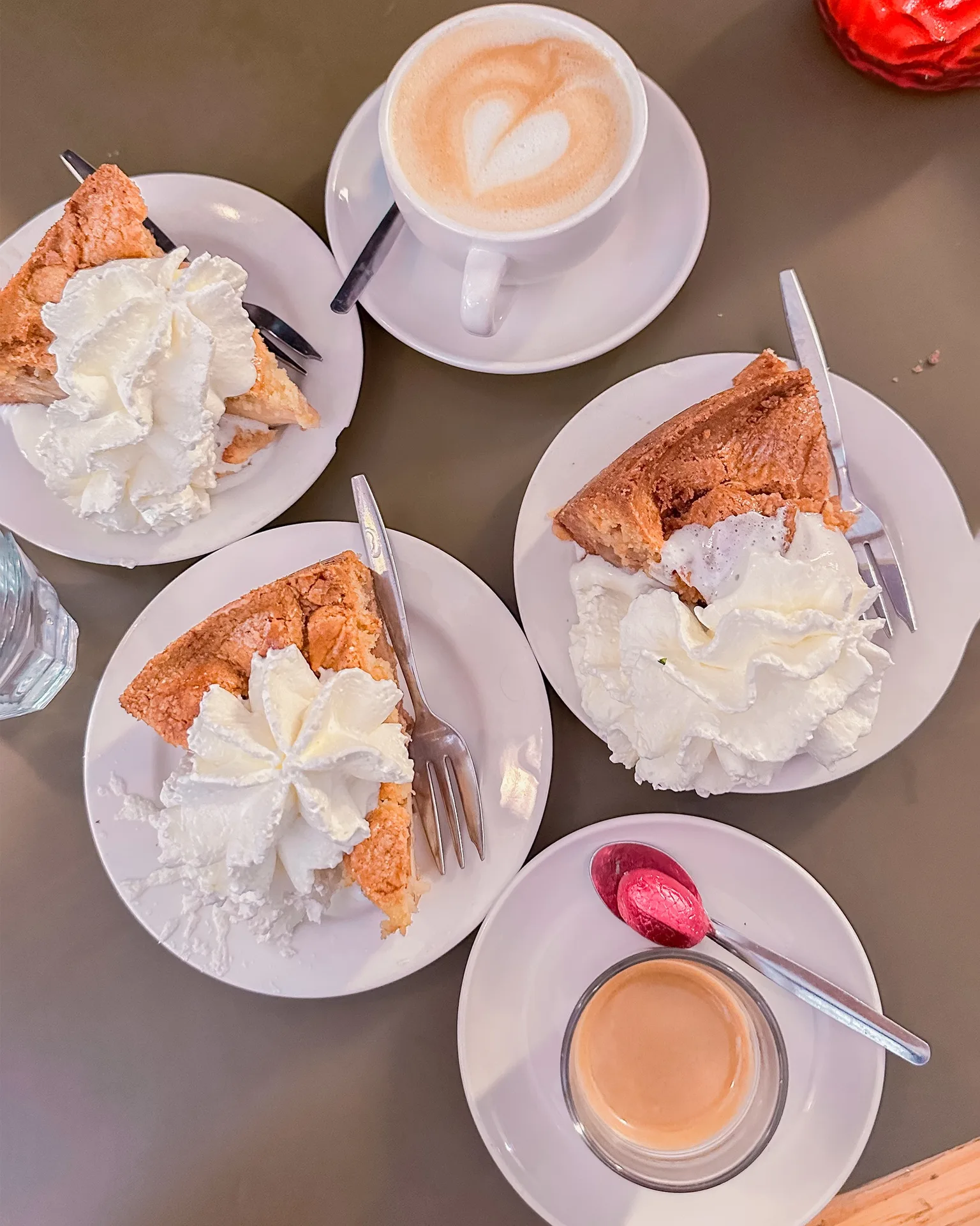 Three plates of cake with generous whipped cream served with a latte and an espresso on a café table.