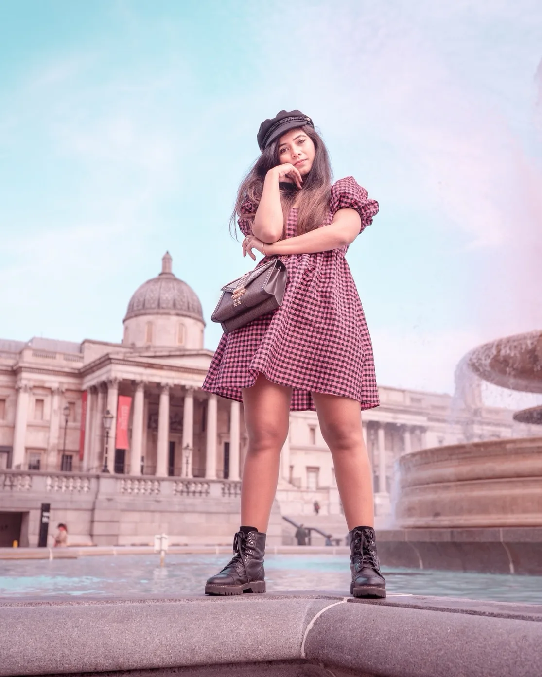 Young woman in a pink gingham dress and black boots posing confidently by a fountain with the National Gallery dome in the background.
