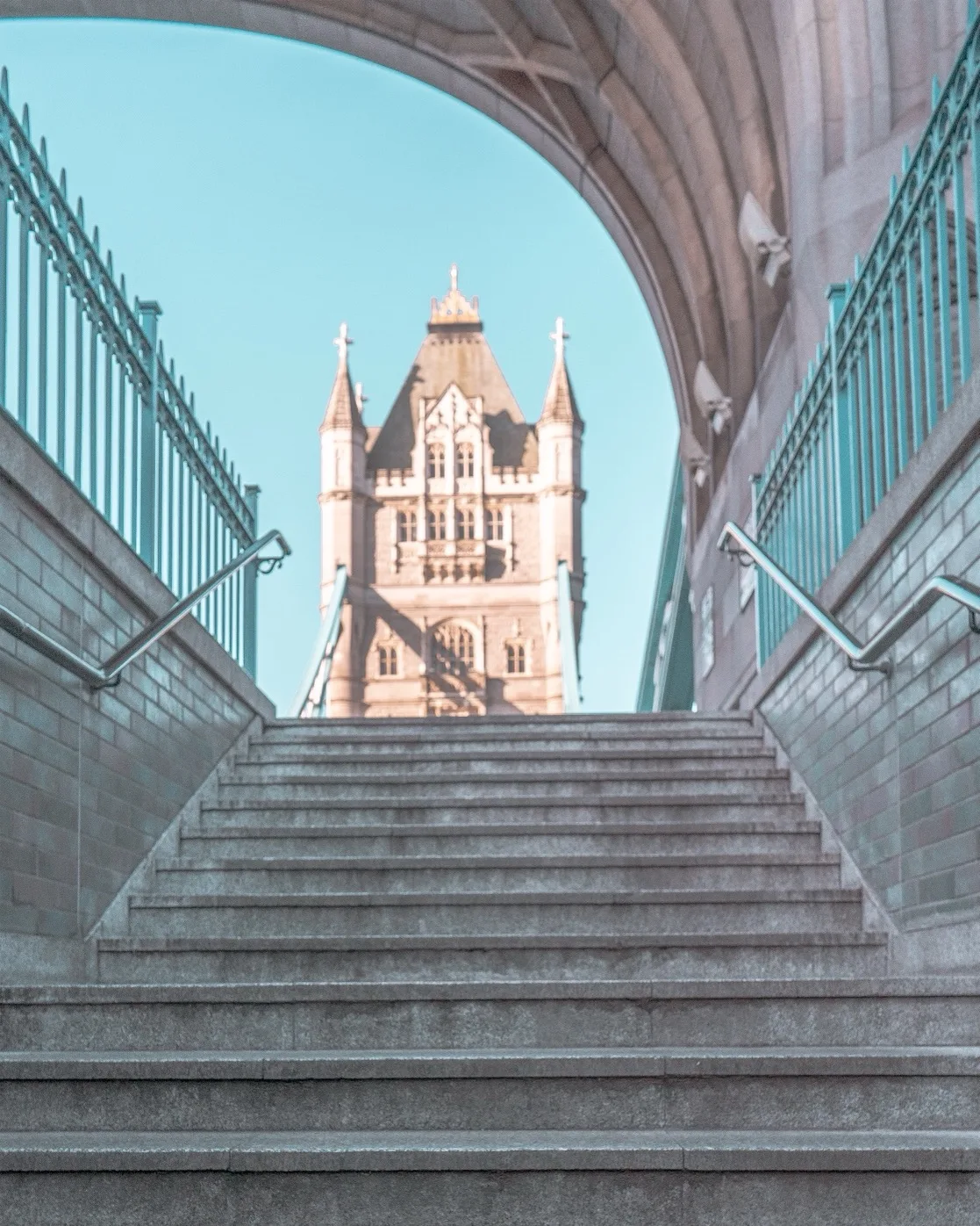 Stone steps leading up through an arched passage to a sunlit ornate tower of Tower Bridge in London.