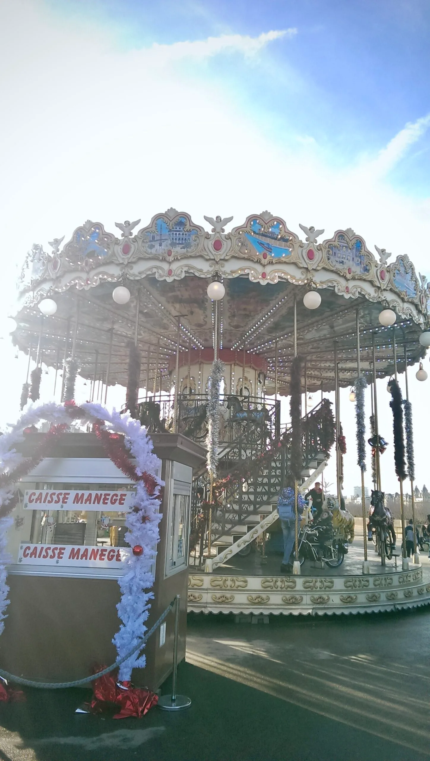 Two-tier vintage carousel decorated with lights and garlands beside a ticket booth labelled "Caisse Manège" under a bright blue sky.