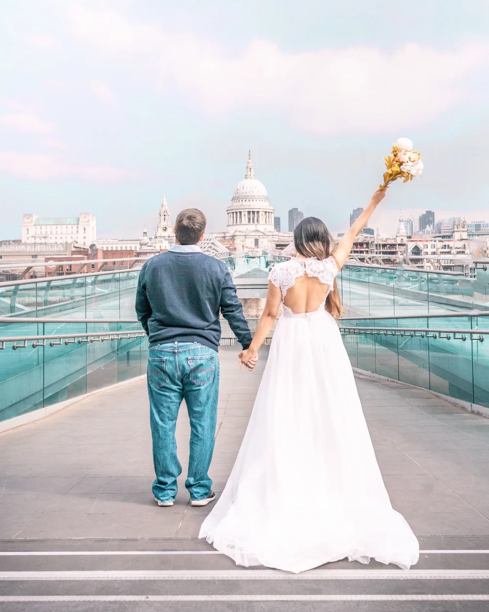 Bride in a white dress holding a bouquet and a casually dressed partner holding hands while facing St Paul’s Cathedral from a glass pedestrian bridge