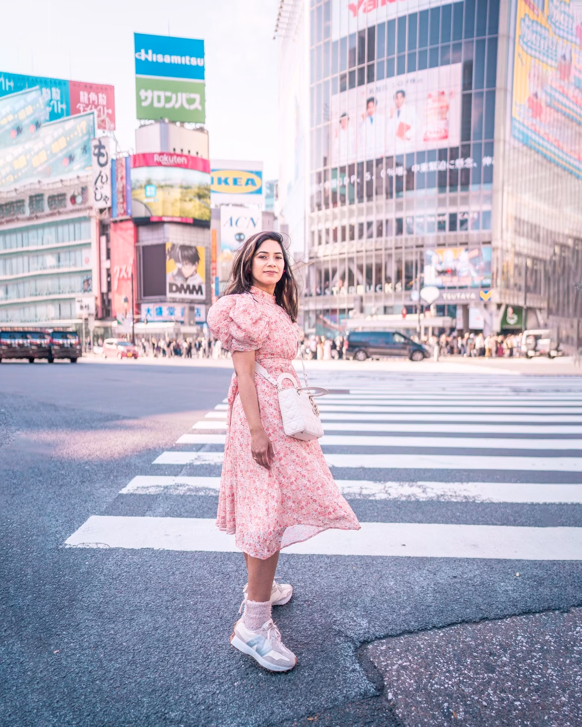 A woman in a pink dress stands on a crosswalk in a busy city district with large billboards and tall buildings