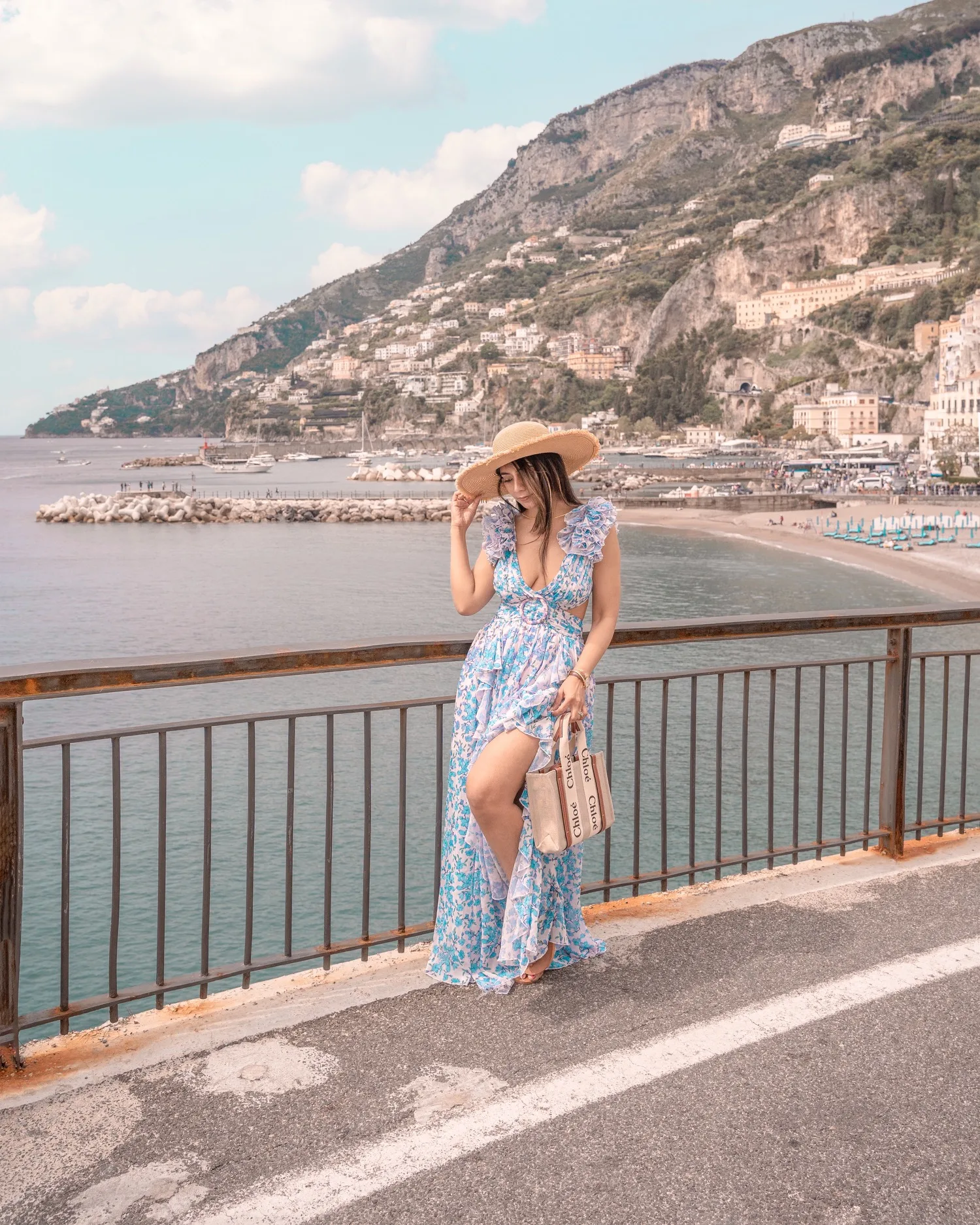Woman in a blue floral sundress and straw hat leaning on a seaside railing with cliffs, villas and a marina in the background.