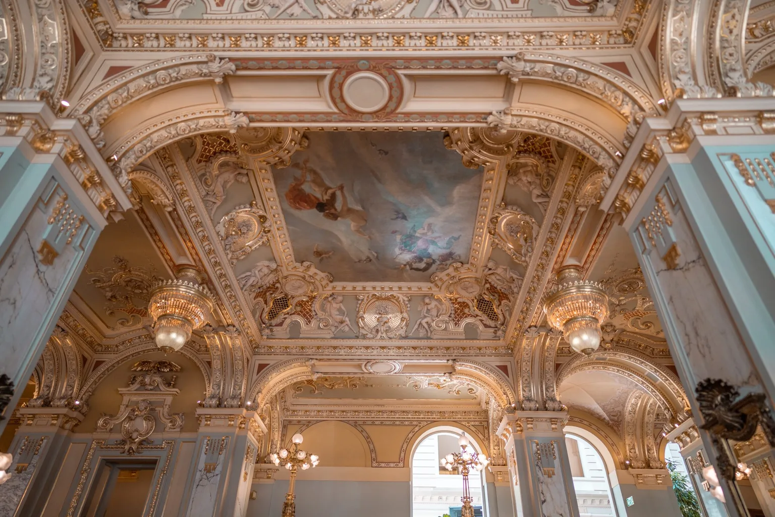 Ornate gilded ballroom ceiling with painted fresco, sculpted mouldings and crystal chandeliers in a grand historic interior.