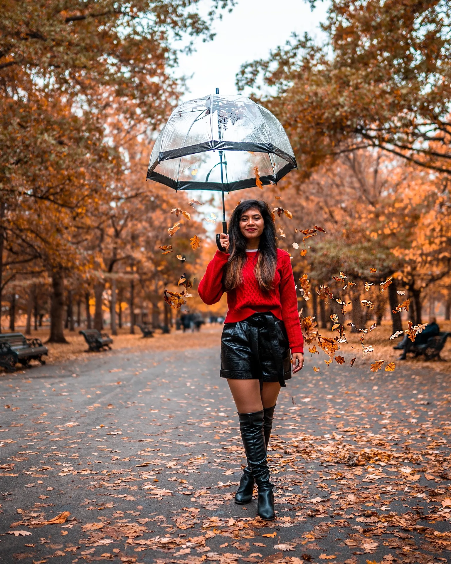 Woman in a red sweater and black leather skirt walking through an autumn park holding a clear umbrella as leaves swirl around her.