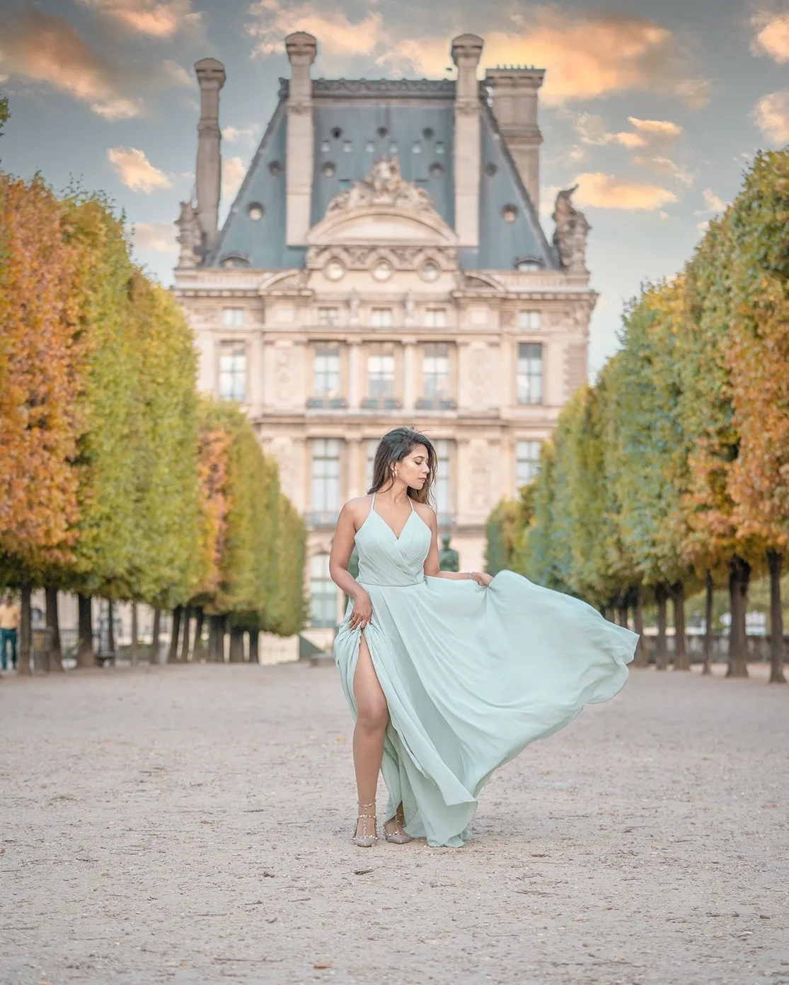 Woman in a flowing pale green gown posing on a tree-lined gravel path with a grand historic museum building in the background.