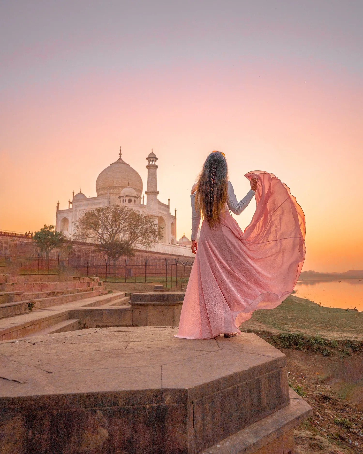 Woman in a flowing pink dress stands on a stone platform gazing at the Taj Mahal at sunrise.